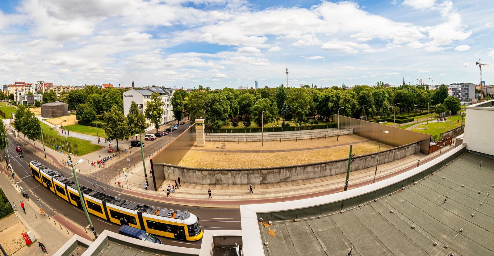 Wide panorama from a rooftop over a preserved section of the Berlin Wall and border strip, with a yellow tram in the foreground and the TV tower on the horizon