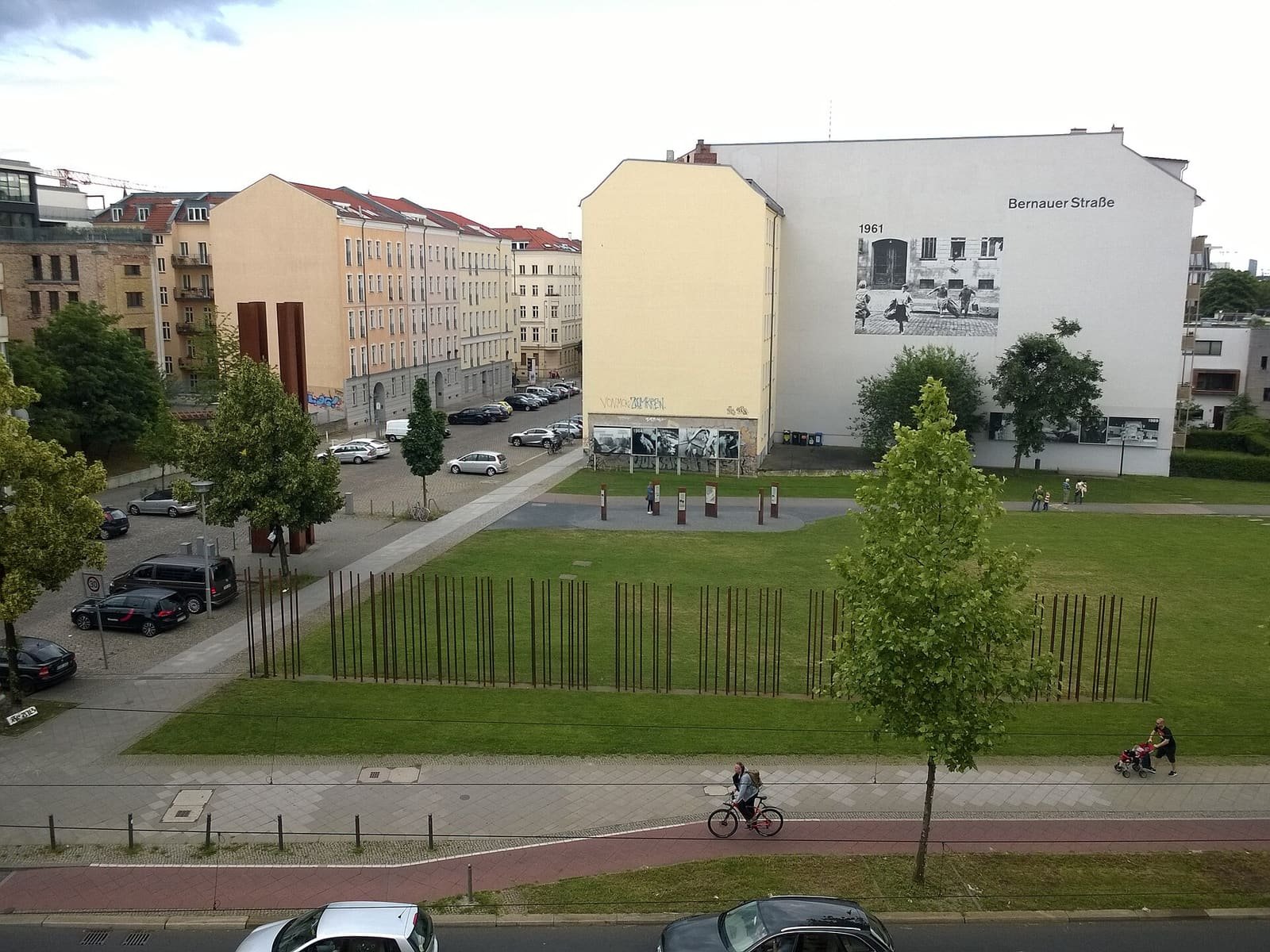 Lawn with rows of rusty steel rods marking the Wall's course, apartment buildings behind with a large black-and-white photo mural