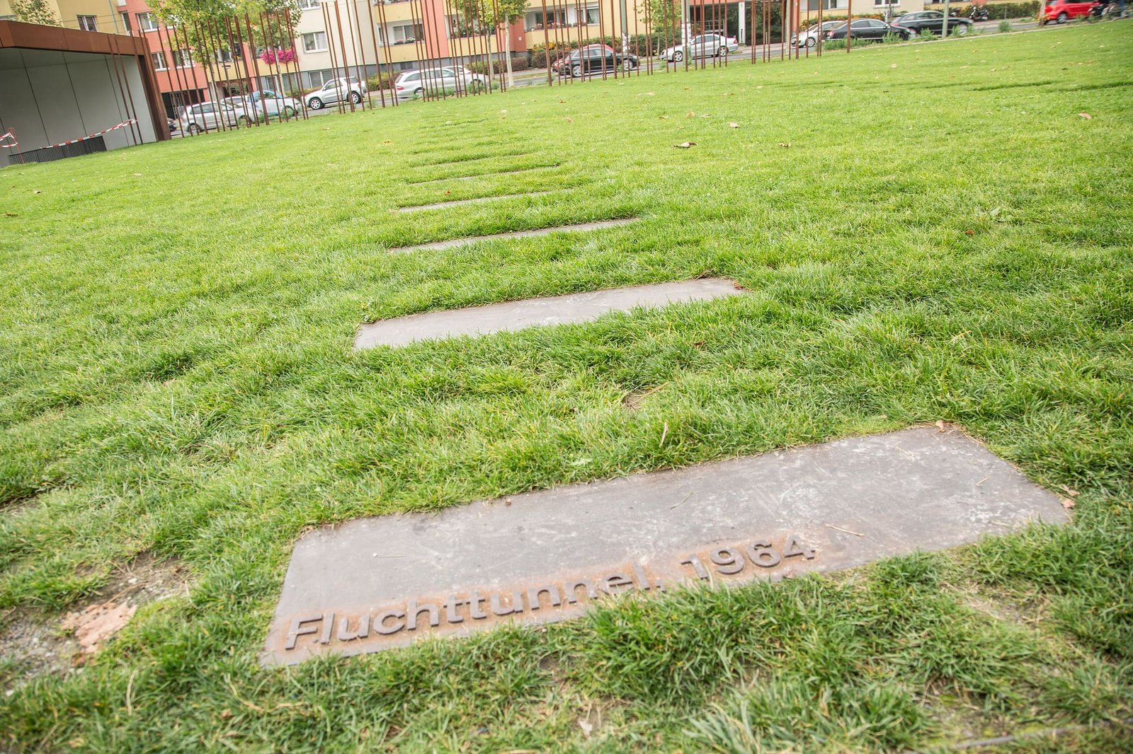A lawn with a line of rectangular steel plates set into the grass; the nearest plate is inscribed 'Fluchttunnel 1964'. Rusty steel rods marking the Wall's course and apartment buildings are behind.