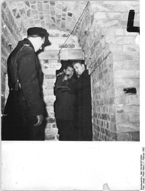 Black-and-white photo of a narrow brick vault. A man in uniform with a shoulder strap stands on the left; two men in coats and hats look out from a low opening in the brickwork, one pointing.