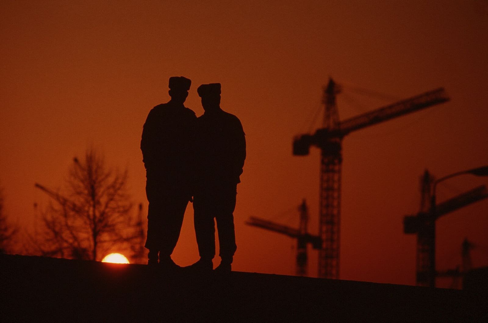 Silhouettes of uniformed men standing on top of the Berlin Wall against an orange sunset sky.