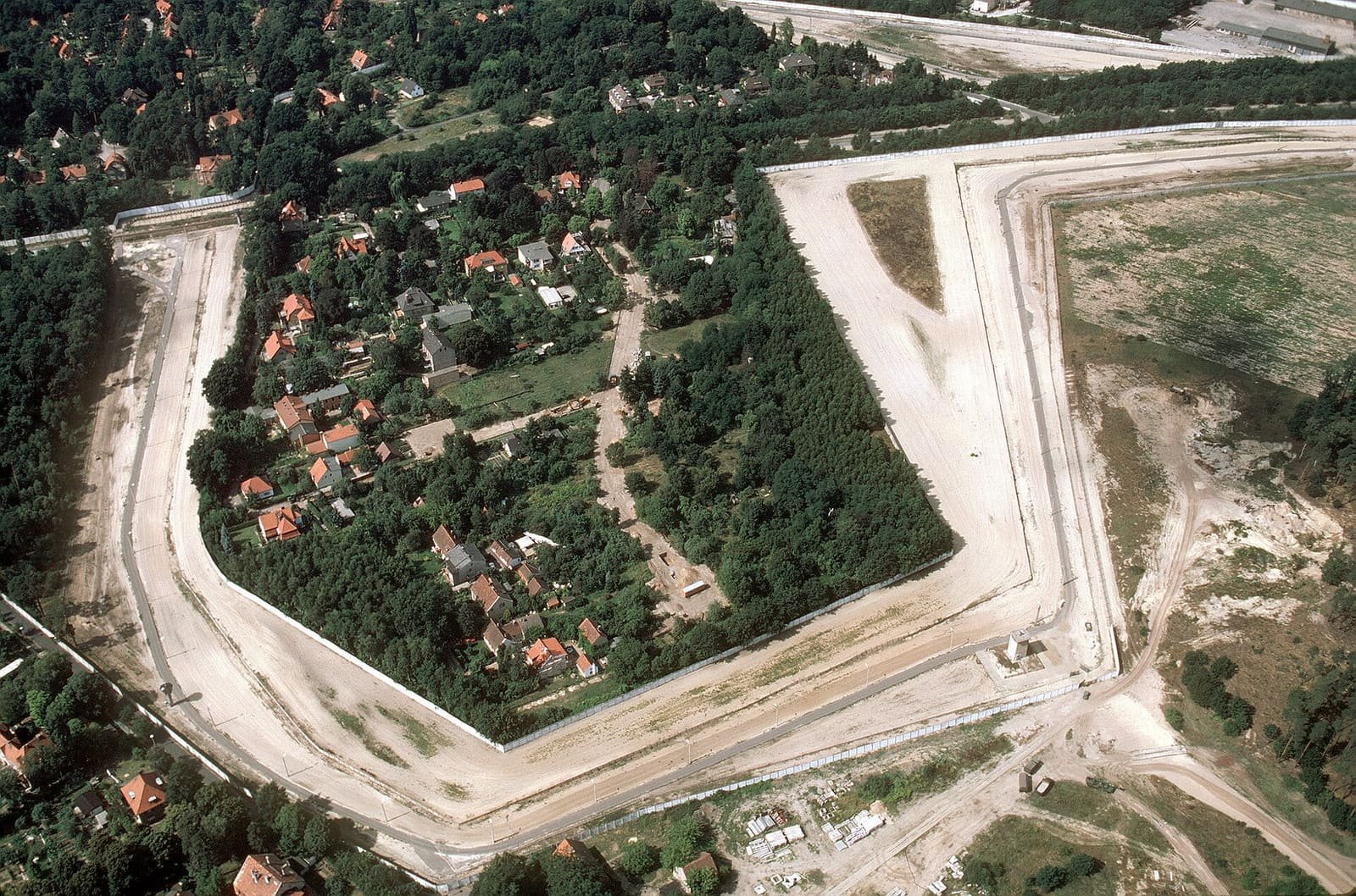 Aerial photo of a wide pale strip of cleared land and a wall bending around a cluster of houses and woodland.