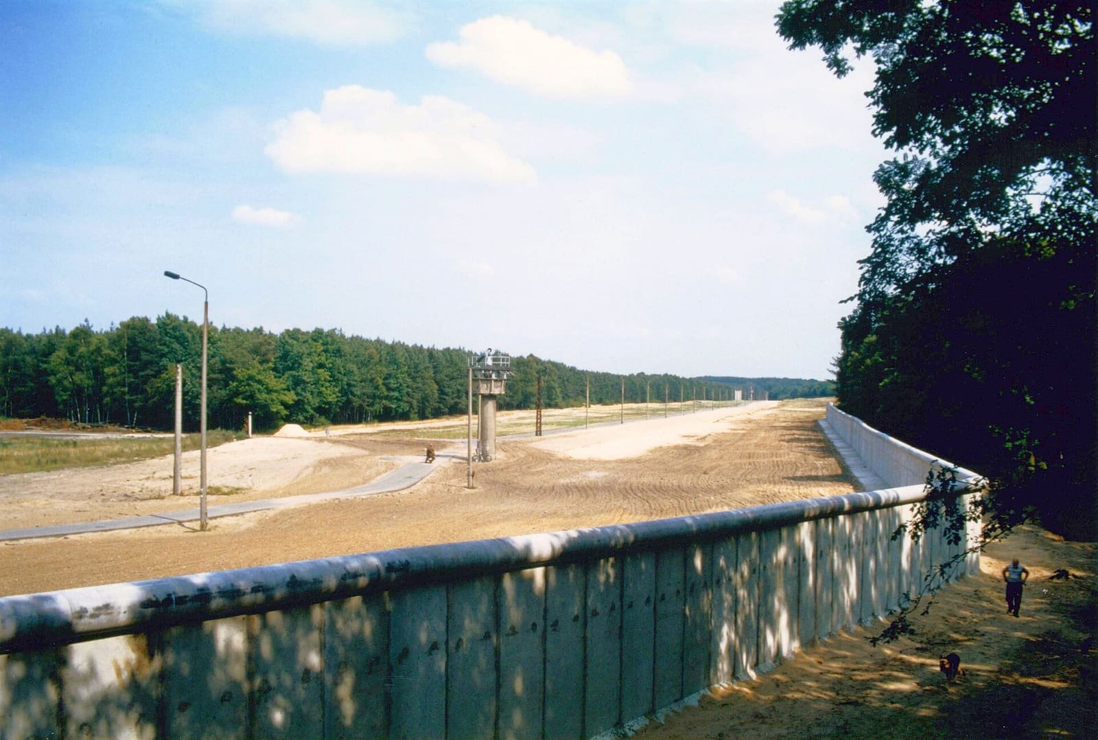 A raked sand strip with a patrol road, lamp posts and a watchtower behind a concrete wall, seen from the West.