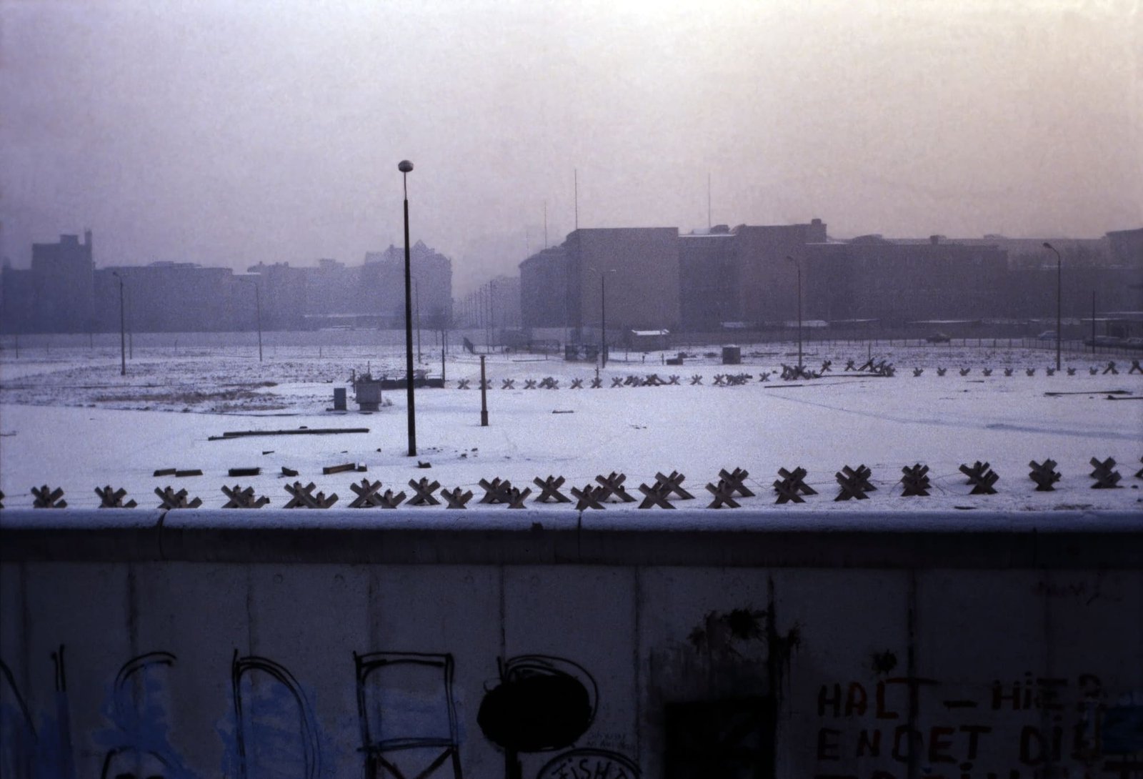 A snow-covered wide strip of ground between two walls, with rows of anti-tank obstacles, lamp posts and a watchtower, seen from above over the top of a concrete wall.