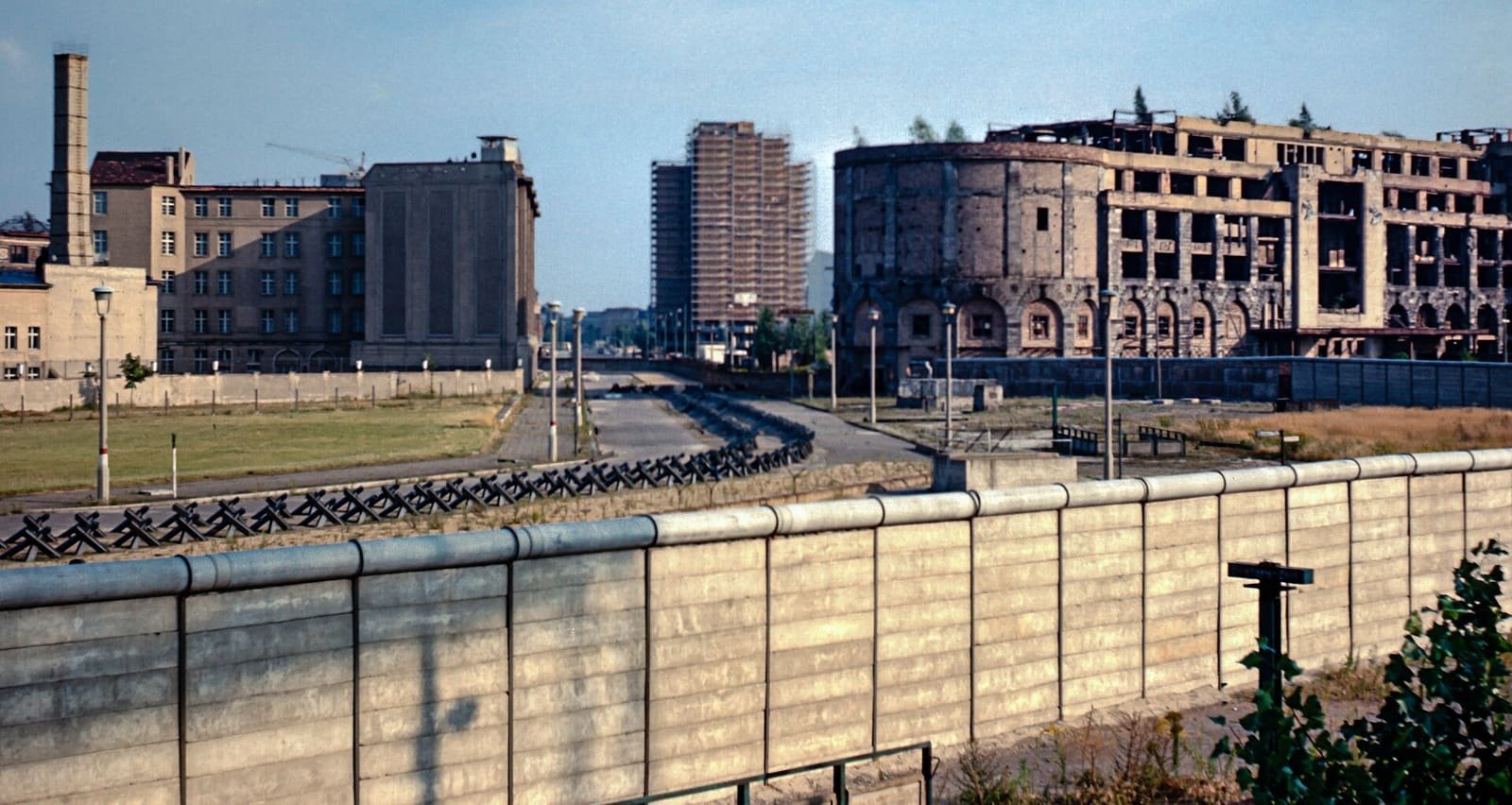A wide view of layered walls, fences and tank traps across an empty square, with a ruined building behind.