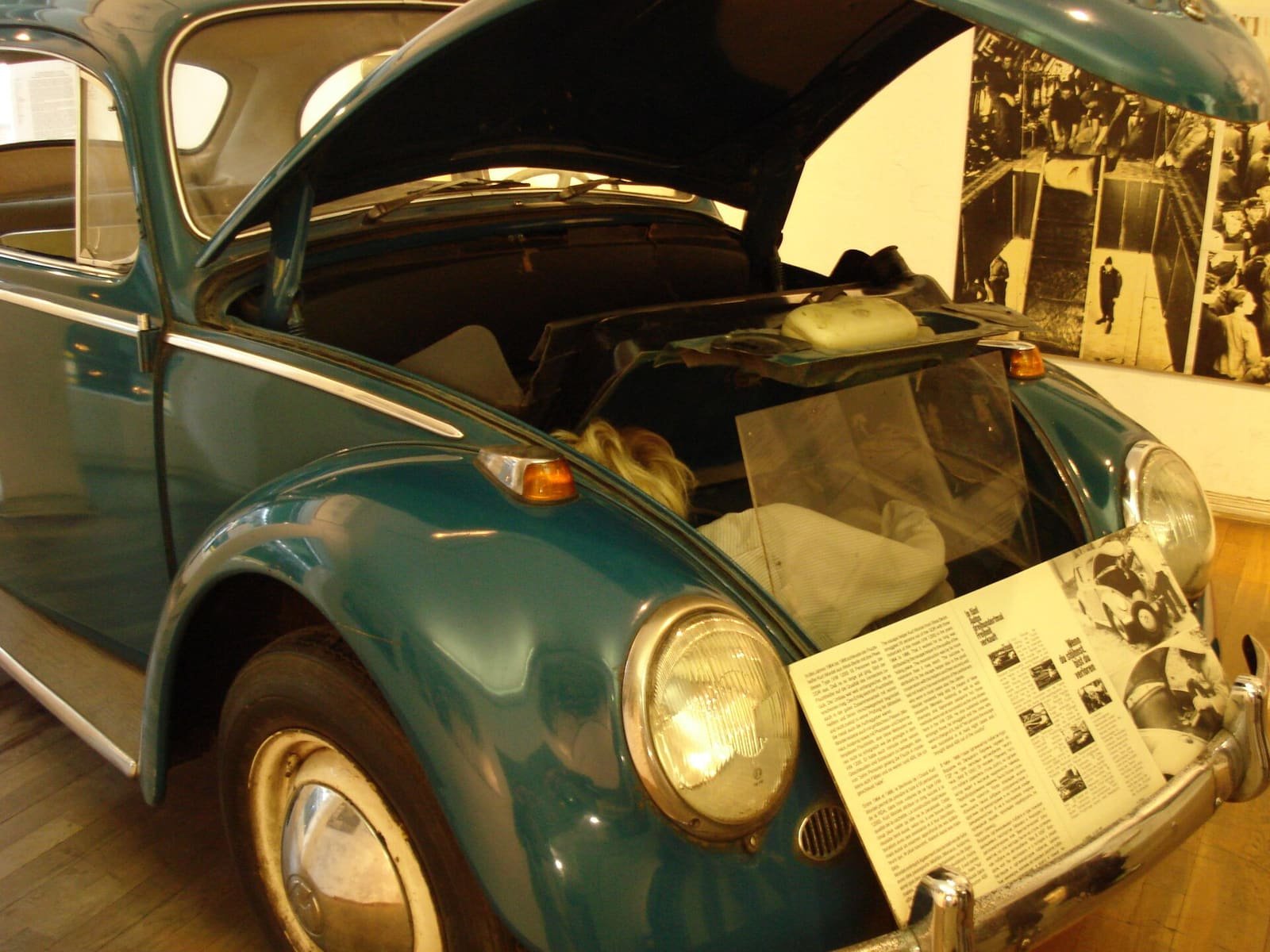 A teal VW Beetle in a museum with its front bonnet open, showing a cramped space, with display boards behind.