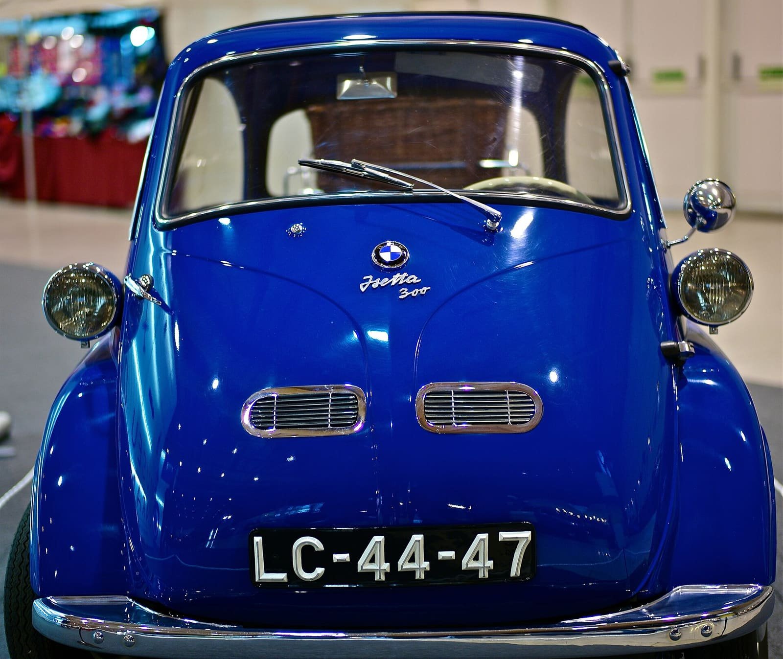 A small, egg-shaped two-tone microcar with a front-opening door, parked at a car show.