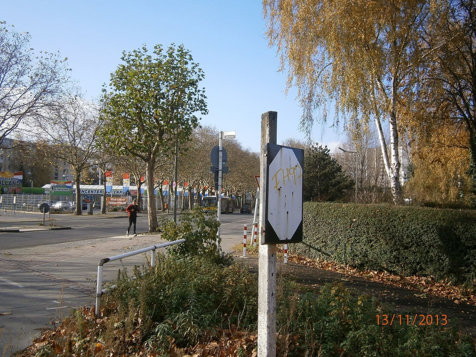Colour photo: an old railway signal post with a white board stands in undergrowth beside a hedge and a wide road; residential blocks and trees are in the background.