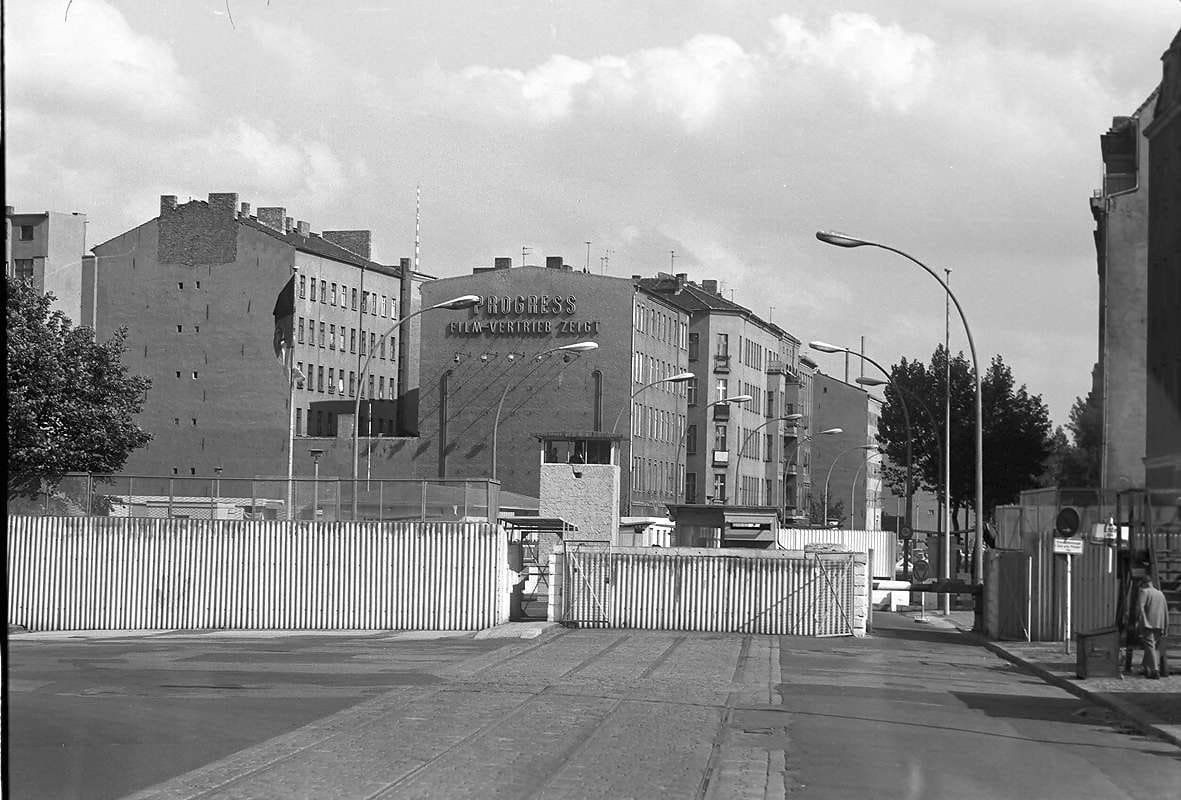 Black-and-white photo of a wide empty street leading to a barrier, the Wall and control buildings at the Chausseestraße crossing, with apartment blocks behind.