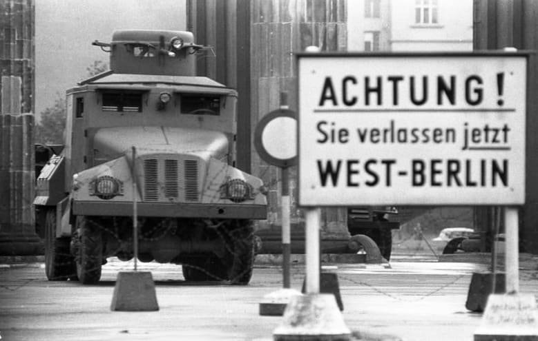 Black-and-white photo: a boxy armoured truck with a turret stands between the columns of the Brandenburg Gate; in the foreground a large sign warns 'Achtung! Sie verlassen jetzt West-Berlin'.