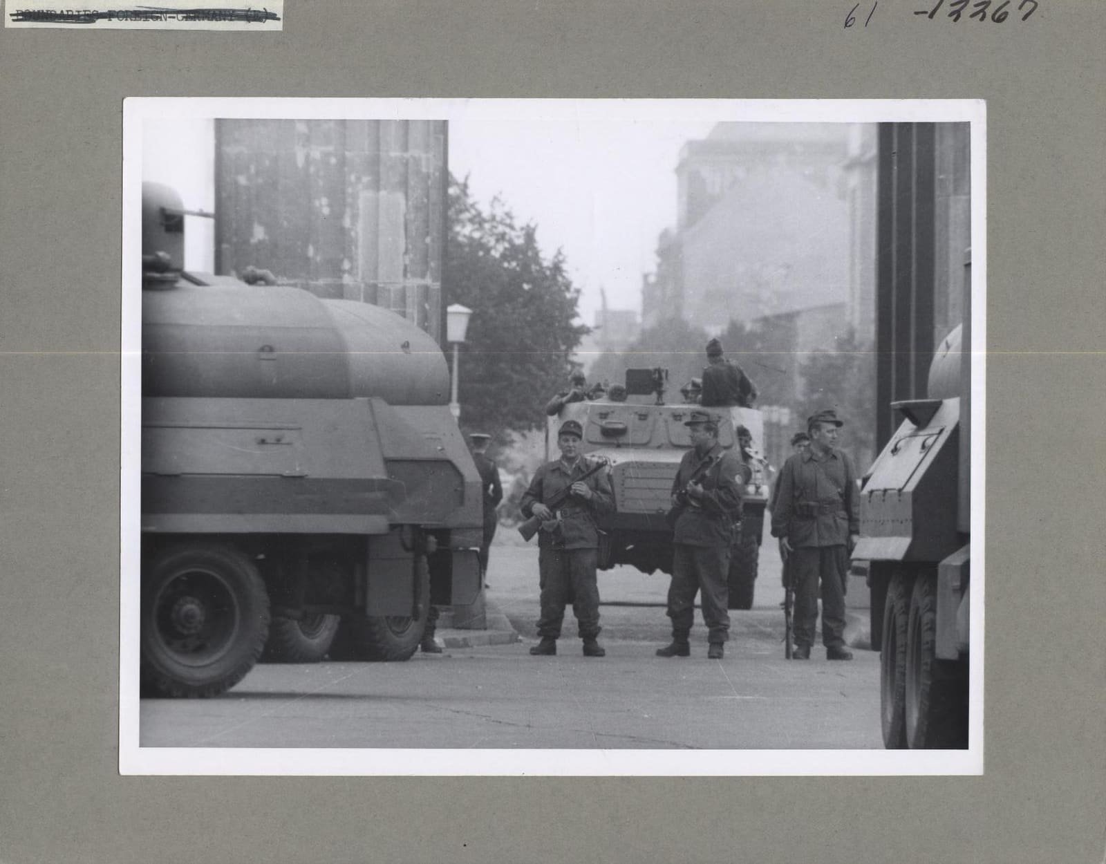 Black-and-white photo: three armed men in uniform stand in front of an armoured car in a passage of the Brandenburg Gate, flanked by large military vehicles.