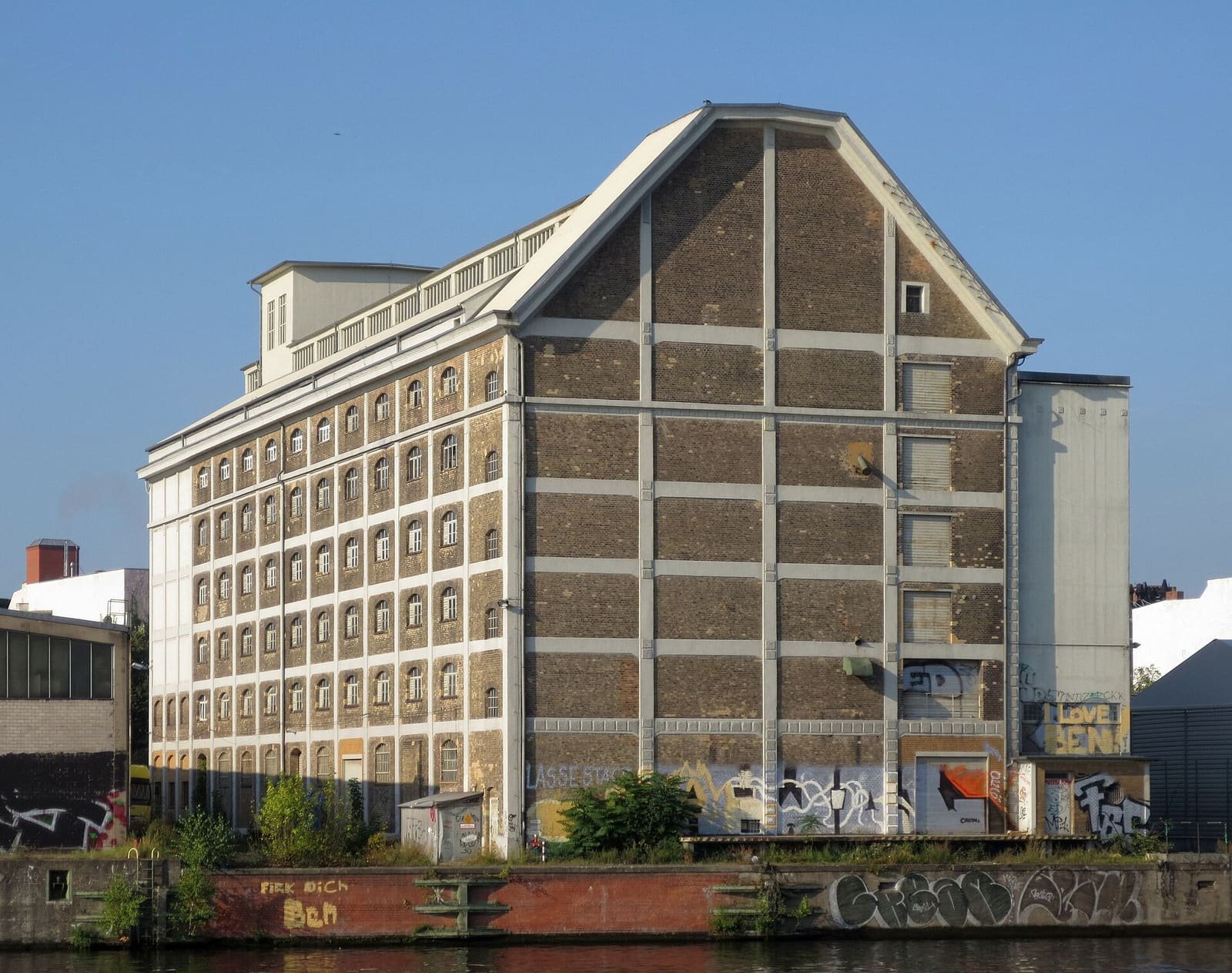 A tall brick warehouse with a pointed gable and rows of small windows on a graffiti-covered riverbank quay.