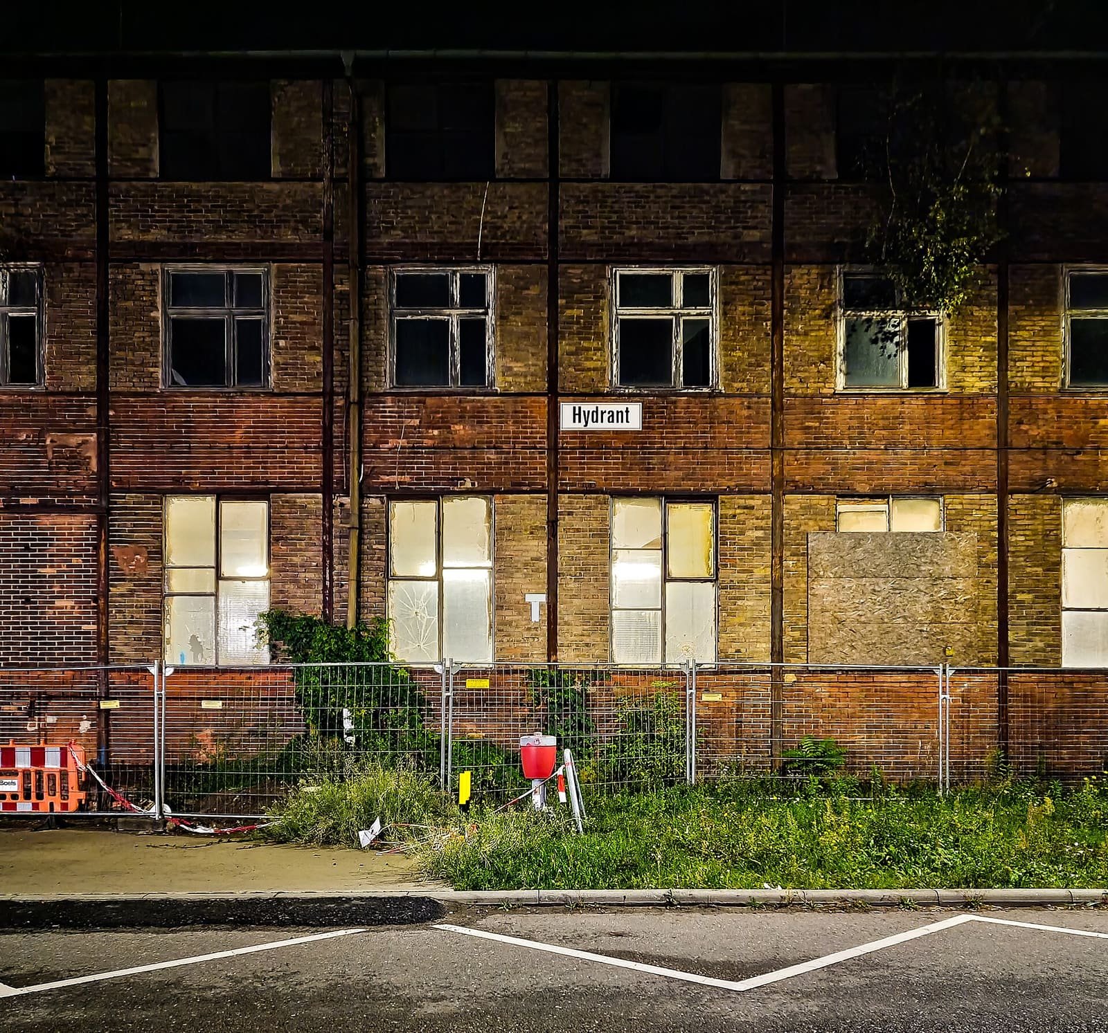 Night view of a long brick industrial building with rows of windows, some boarded up, behind a construction fence.
