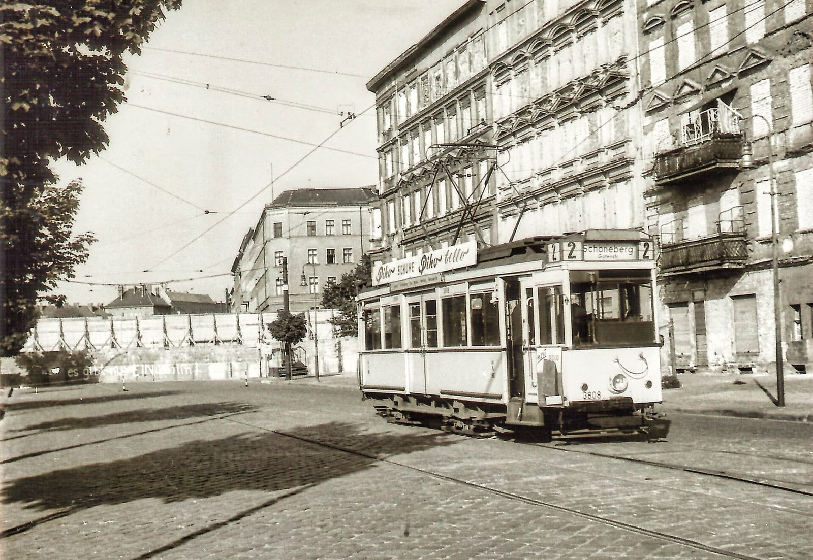 Sepia photo of an old tram on a cobbled street, with tall tenement houses whose ground-floor windows are bricked up on the right and a screen wall across the street behind.