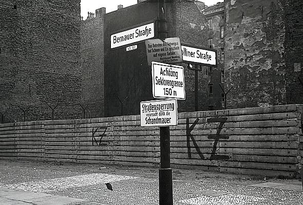 Black-and-white photo: a signpost with street signs for Bernauer Straße and Wolliner Straße and notices reading 'Achtung Sektorengrenze 150 m' and 'Straßensperrung verursacht durch die Schandmauer' stands in front of a low wall of concrete slabs daubed 'KZ', with tall, bare-walled buildings behind.