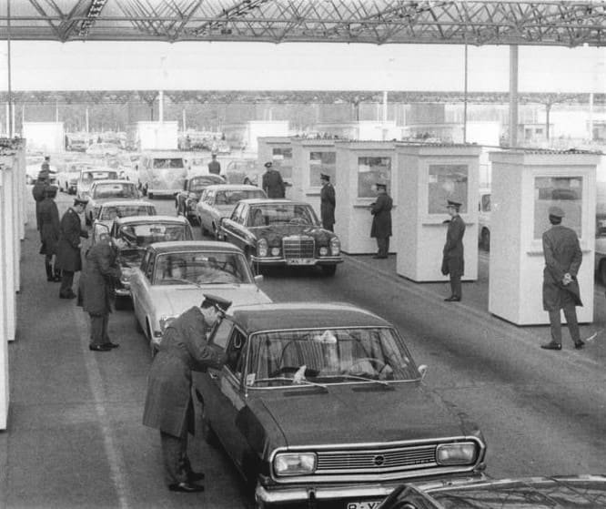 Black-and-white photo: rows of cars queue between white control booths under a large steel canopy; uniformed officers stand at the booths and one bends to speak to the driver of an Opel in the foreground.