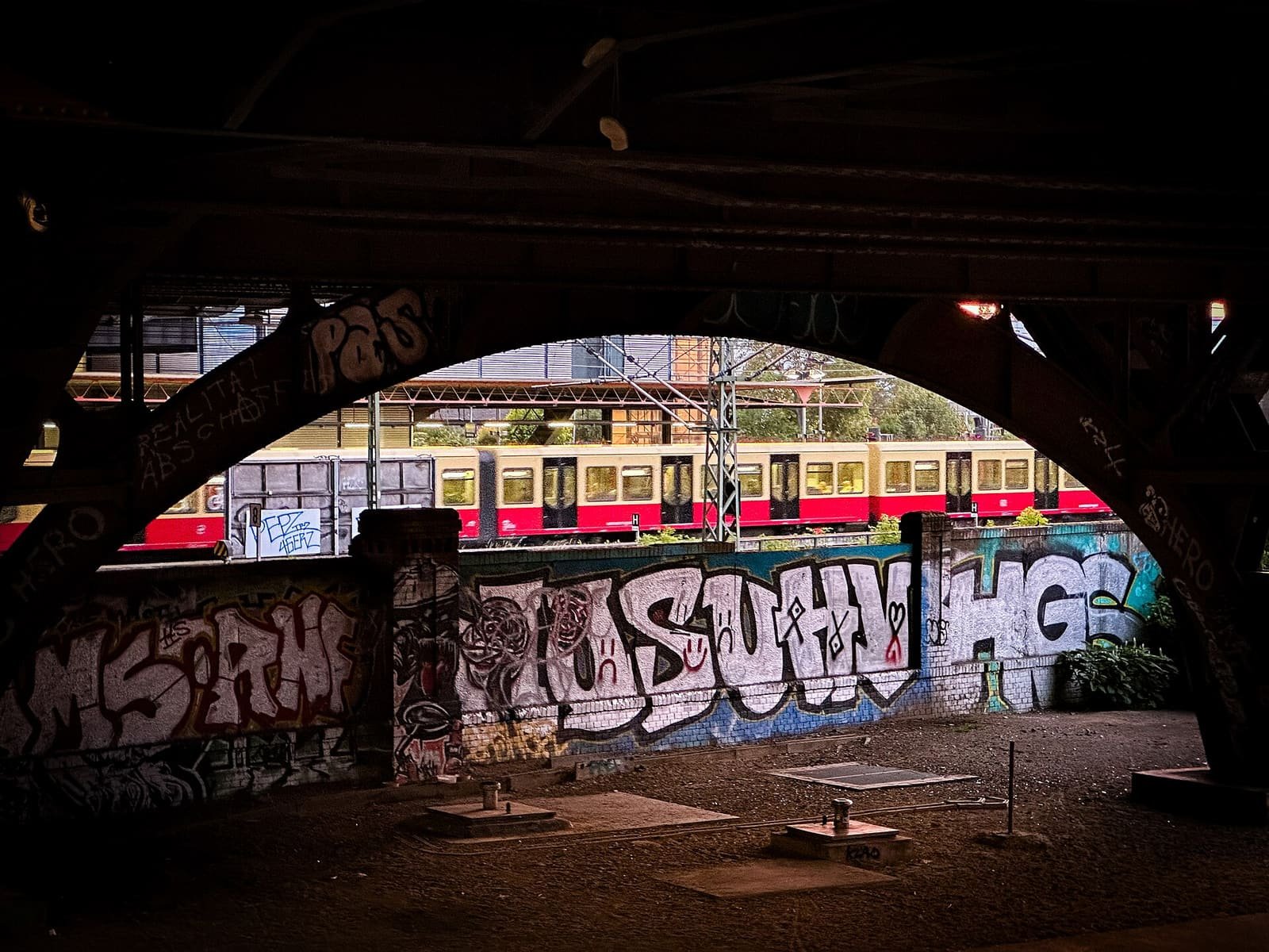 View from under dark steel bridge arches towards a red and cream S-Bahn train passing behind a graffiti-covered wall.