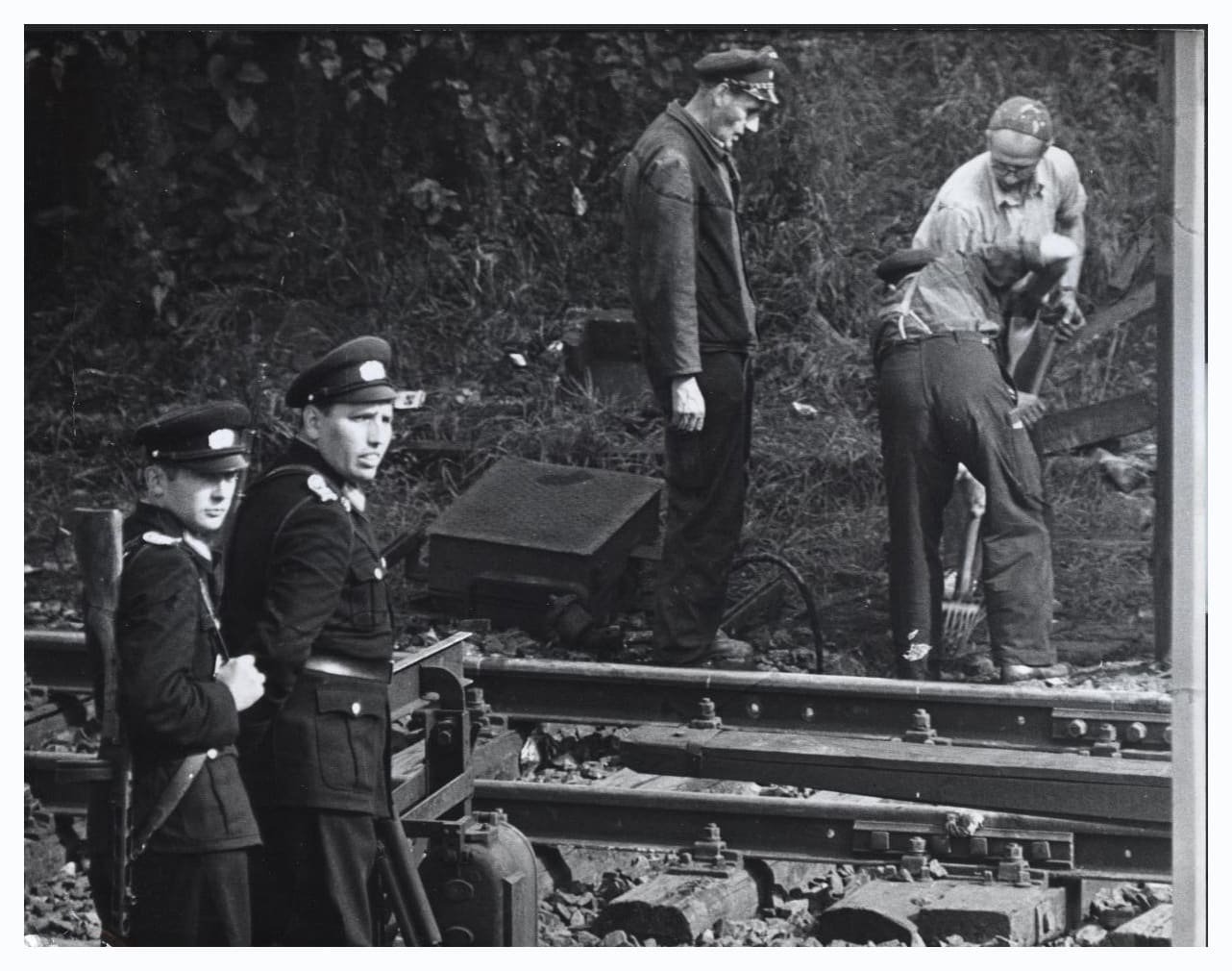 Black-and-white photo: two East German railway policemen in peaked caps stand guard on the left while workmen with pickaxes take up the rails of a railway track beside an overgrown embankment.