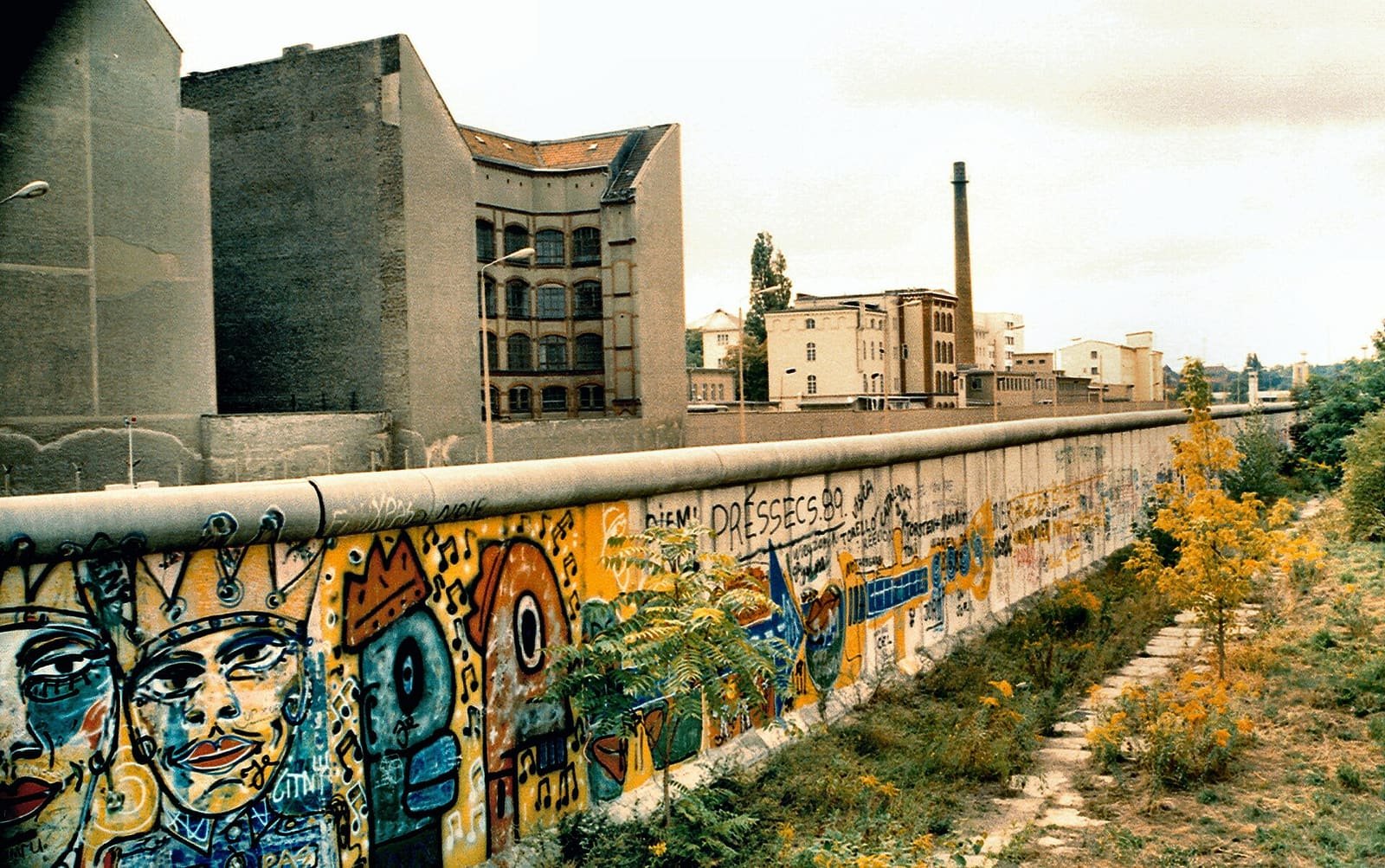 A painted Berlin Wall running along a path with weeds, with old East Berlin tenement and factory buildings and a tall chimney behind it.