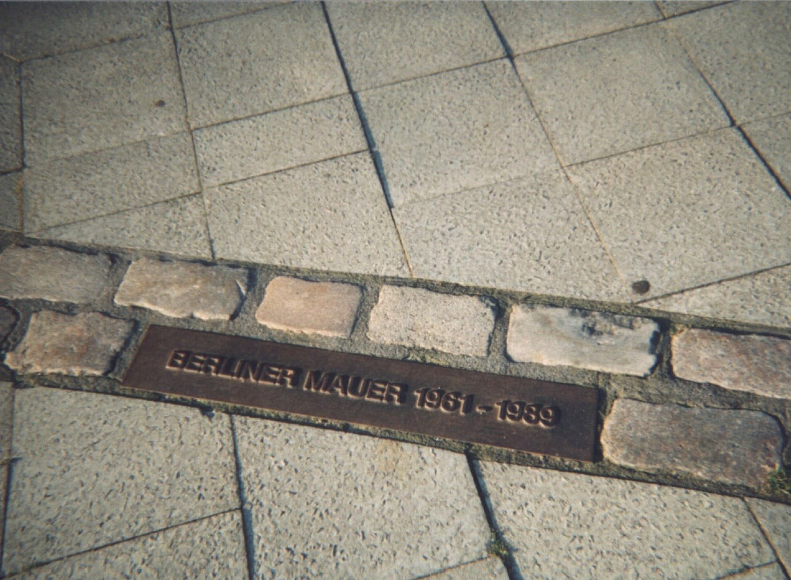 A double row of cobblestones set into paving with a metal plate reading 'Berliner Mauer 1961–1989'.