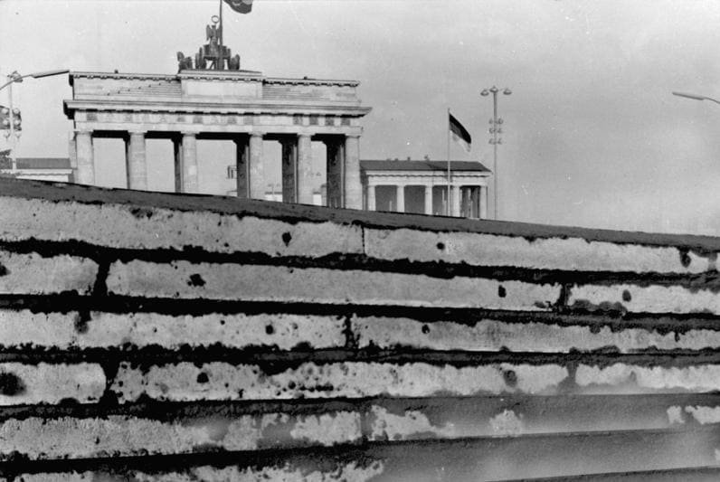 Black-and-white photo of a rough concrete wall in the foreground with the Brandenburg Gate and an East German flag rising behind it.