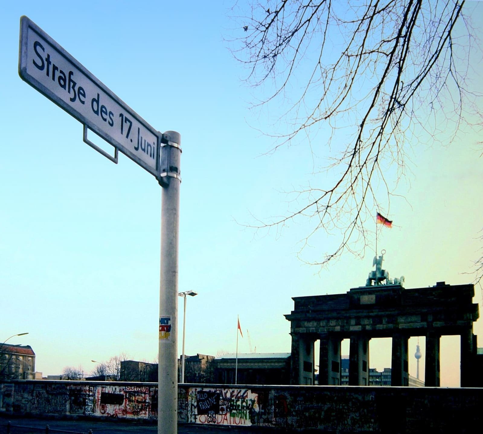 Colour photo: a street sign reading 'Straße des 17. Juni' in the foreground, and behind a graffiti-covered wall the Brandenburg Gate with a flag flying above it and the Television Tower in the distance, under bare branches.