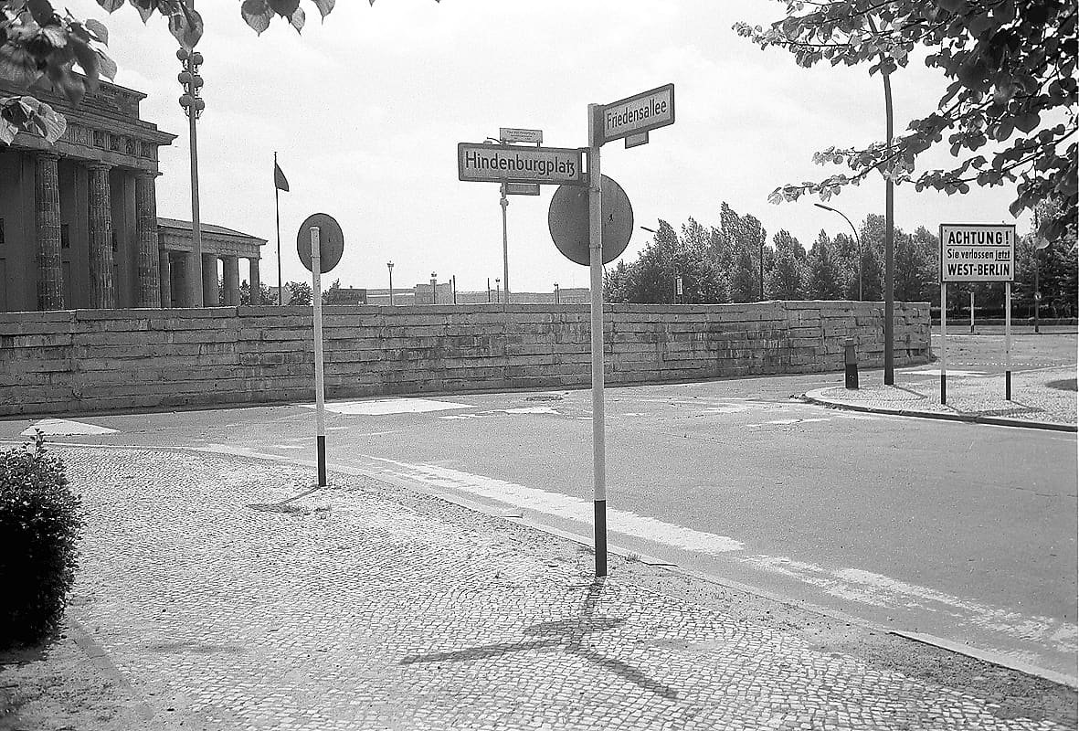 Black-and-white photo of a low, thick wall of stone slabs in front of the Brandenburg Gate's columns, with street signs and a sign reading 'Achtung! Sie verlassen jetzt West-Berlin' in the foreground.