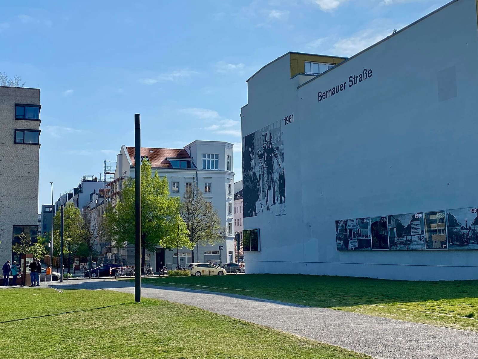 Sunny street corner with a grass strip and path in front, low apartment buildings behind, and on the right a white gable wall labelled 'Bernauer Straße' with a large black-and-white mural and a row of photo panels.