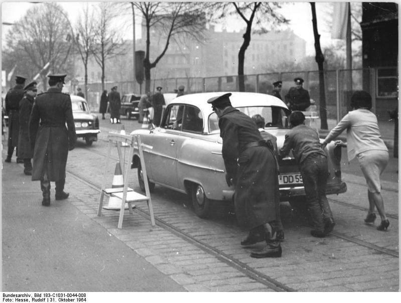 Black-and-white photo of uniformed guards and civilians pushing a car past a small barrier at a checkpoint on a tree-lined street.