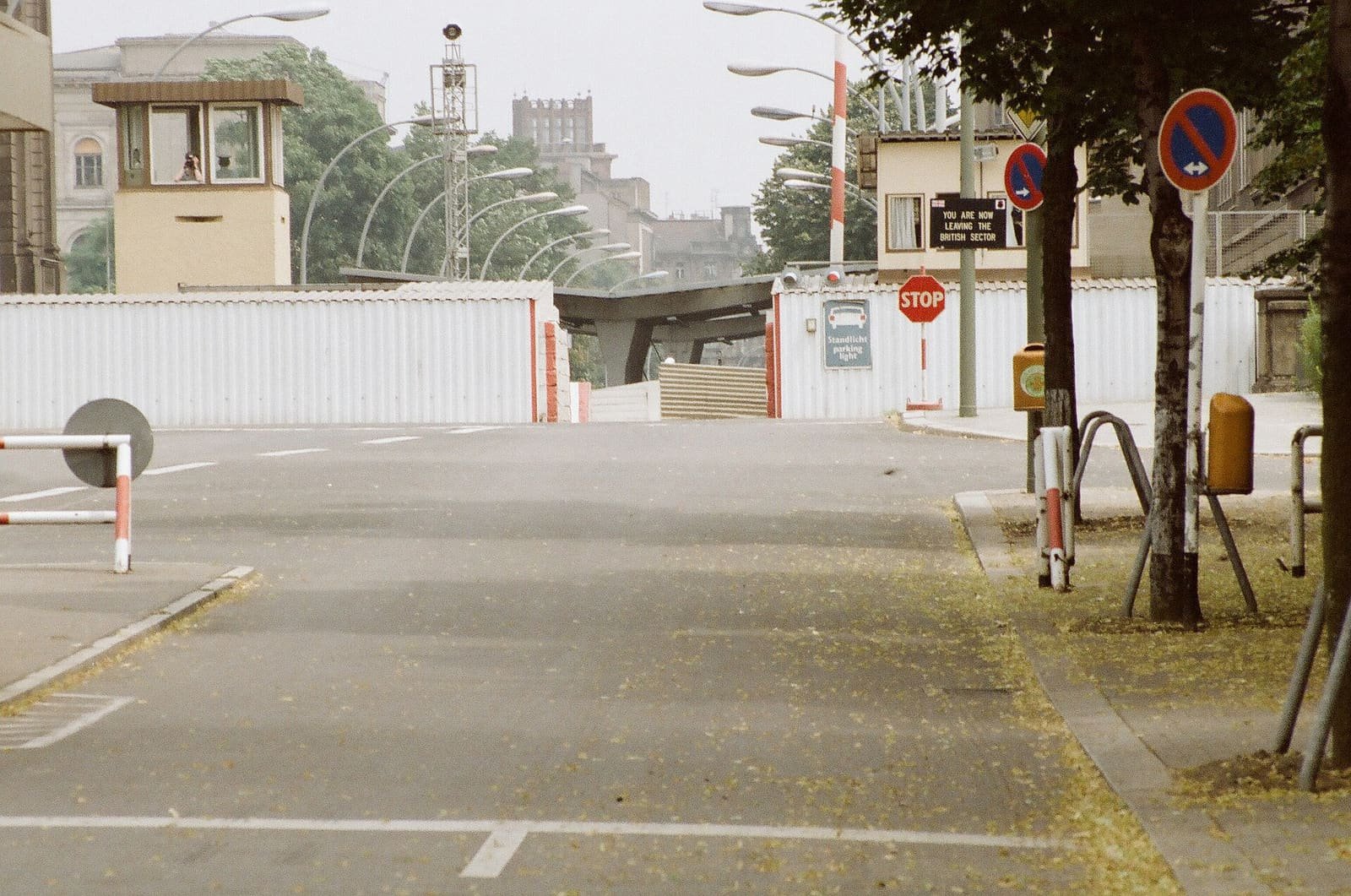 A quiet street leading to a checkpoint with white screen walls, a watchtower, floodlights and a sign reading 'You are now leaving the British sector'.