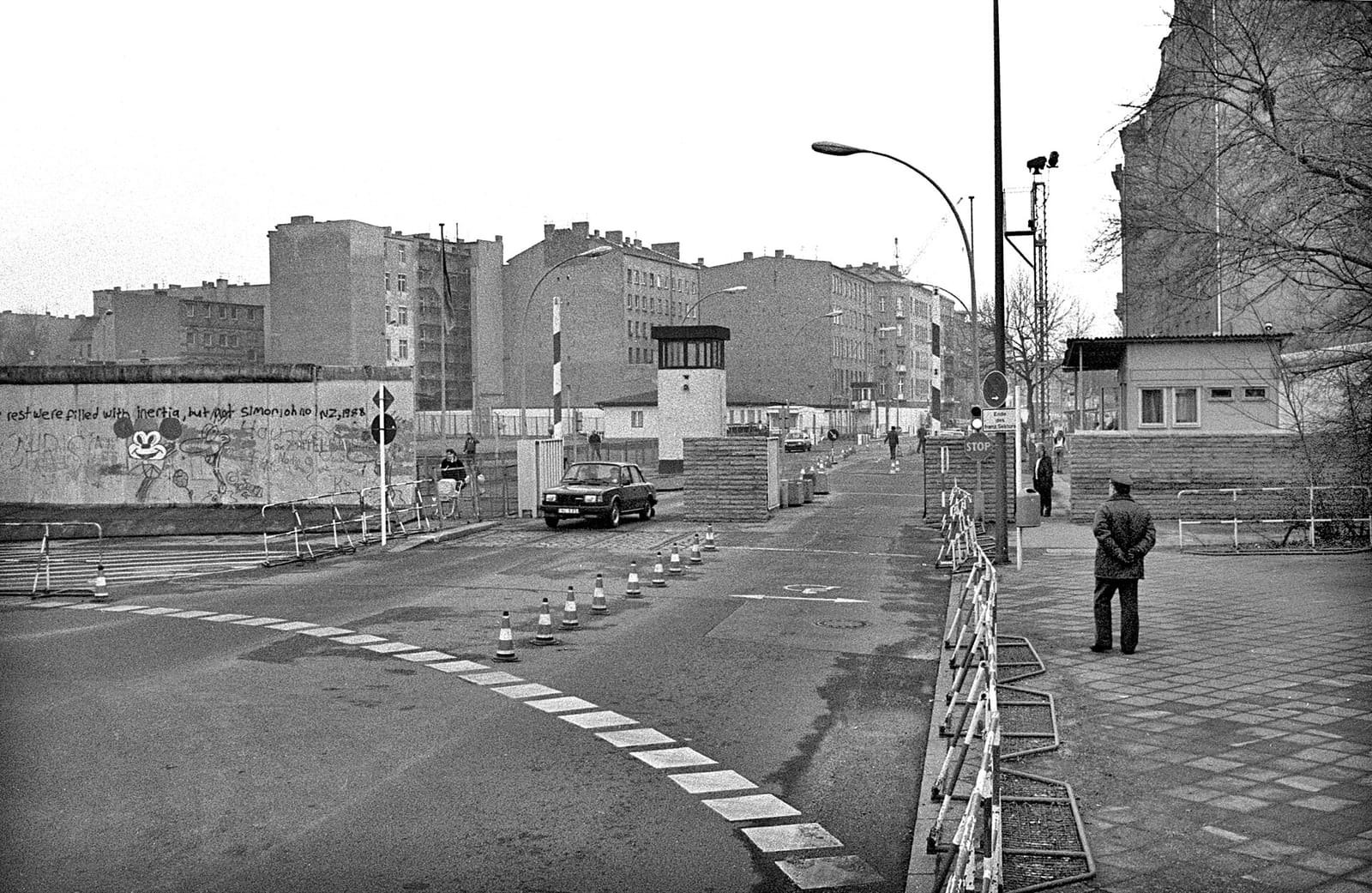 Black-and-white view of a street leading through a border crossing, with traffic cones, a watchtower, a guard hut and a painted wall on the left.