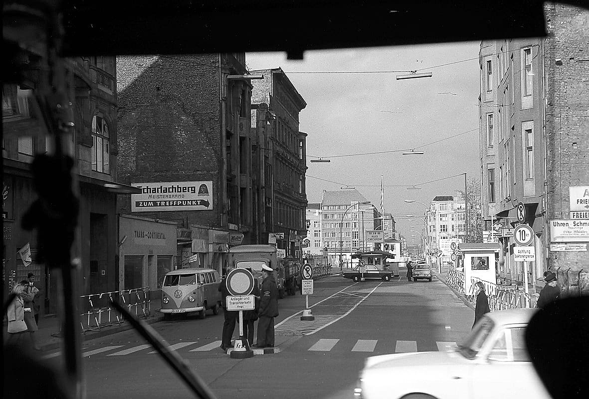 Black-and-white photo through a vehicle windscreen of a street with a VW bus, a traffic island with a man in uniform, barriers and a guard hut in the distance.