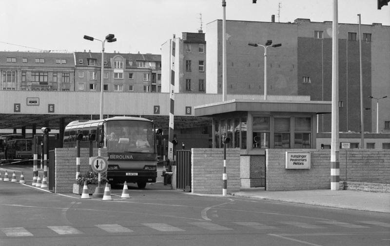 Black-and-white photo of a coach entering numbered control lanes under a canopy, with a gatehouse, brick walls and a sign reading 'Fußgänger Pedestrians Piétons'.