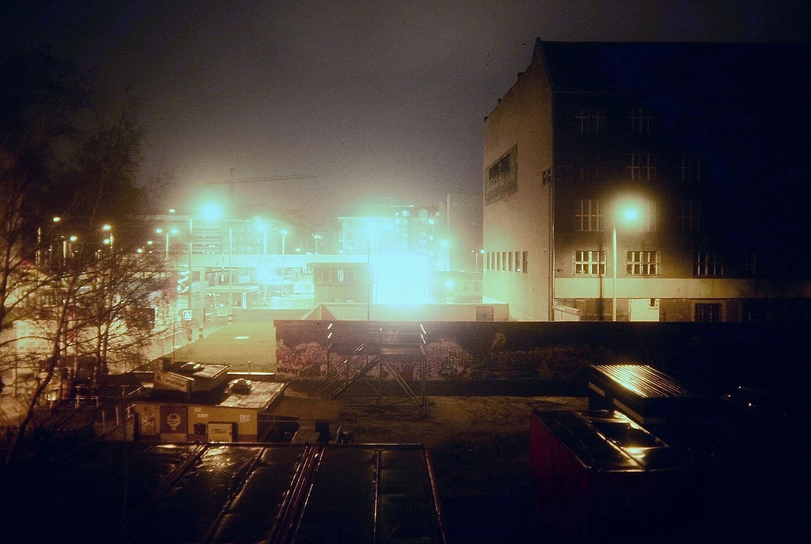 Night photo looking over a graffiti-covered wall into a brightly floodlit control area, with a dark building at right and street lights glowing in mist.