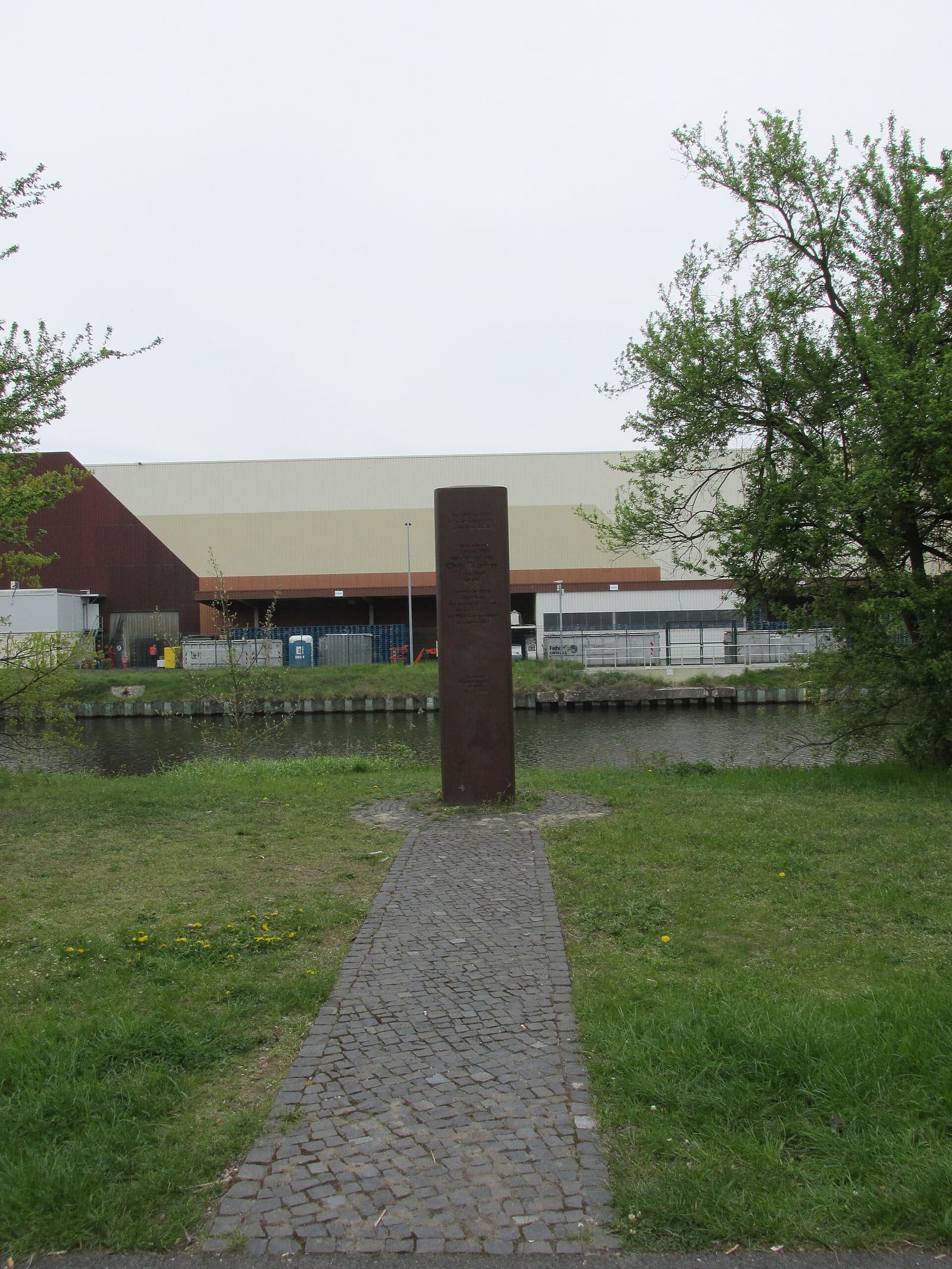 Steel memorial steles seen from a distance on a canal bank, with trees and water beyond