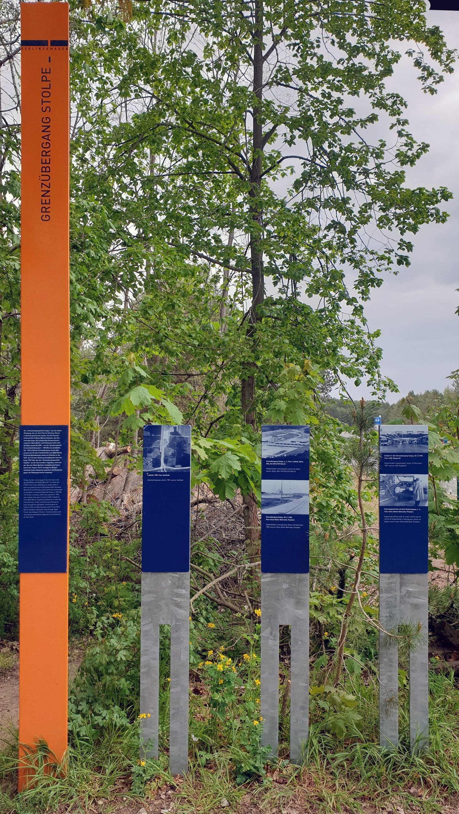 A tall orange stele reading 'Grenzübergang Stolpe' and three blue panels with photos, among trees.