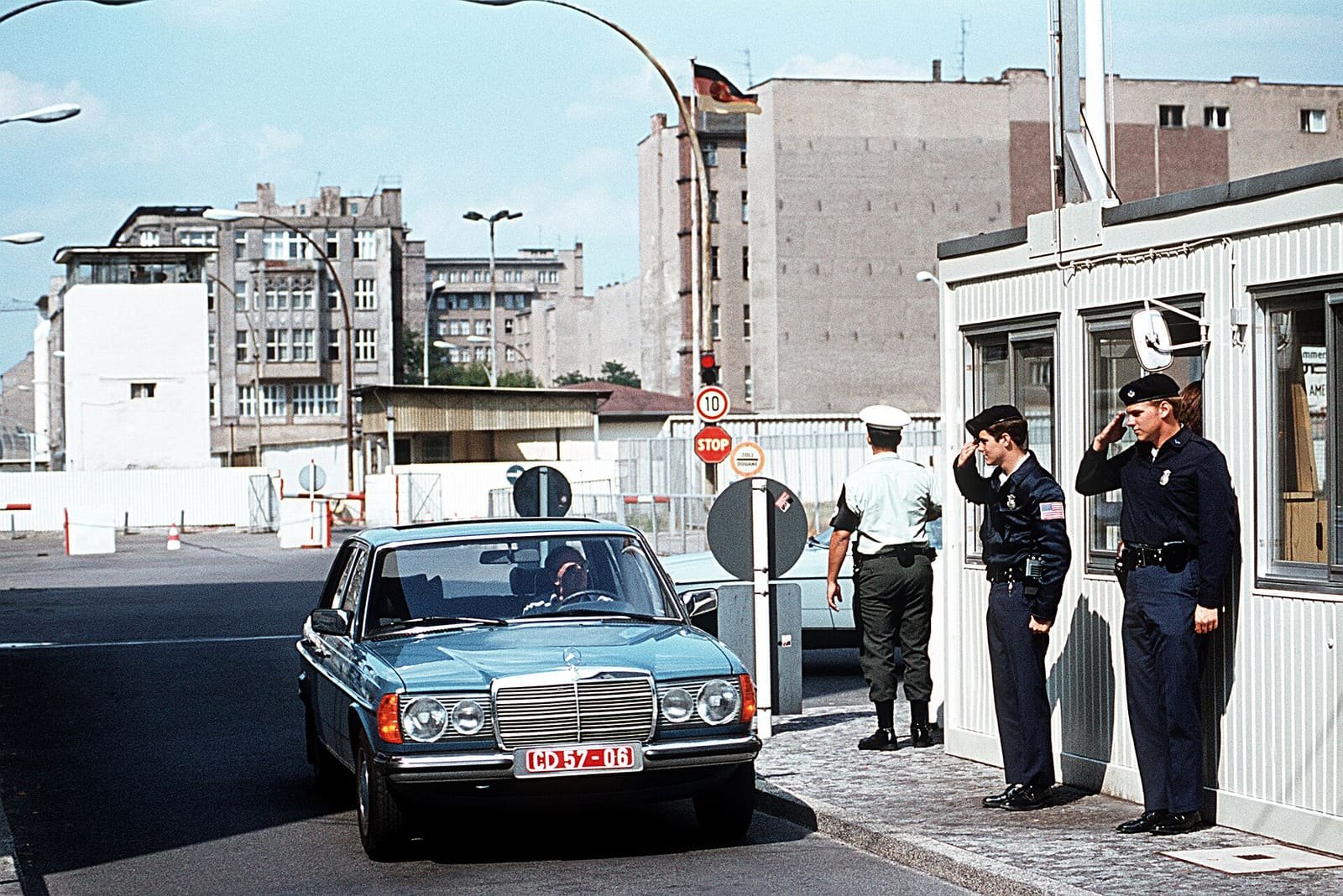 Colour photo of a blue Mercedes saloon with 'CD' plates at a checkpoint; two uniformed US policemen salute beside a white hut, with a GDR flag and buildings behind.