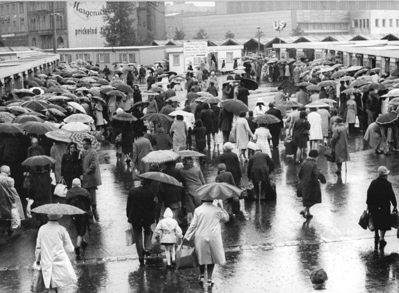 Black-and-white photo: a dense crowd of people with umbrellas and bags walks across a wet square in front of low pavilion buildings, with a large advertising hoarding on a building behind.