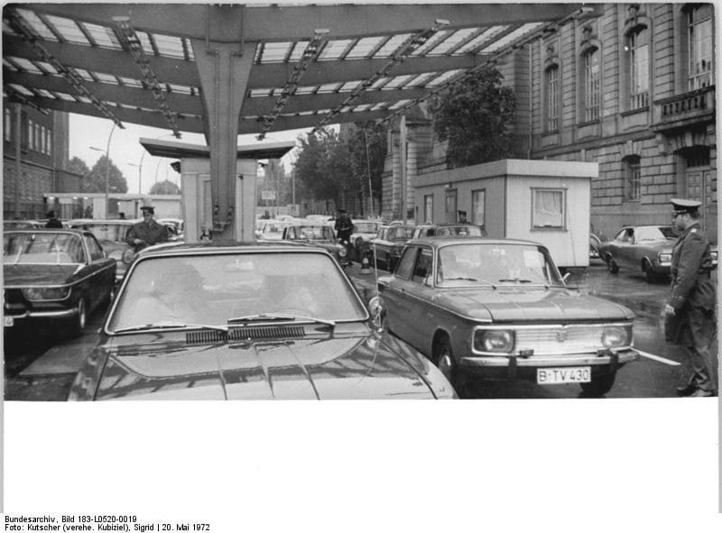 Black-and-white photo: cars, including an NSU with a Berlin number plate, wait in lanes beneath a ribbed canopy beside a small control hut; uniformed officers stand between the lanes in front of a large stone building.