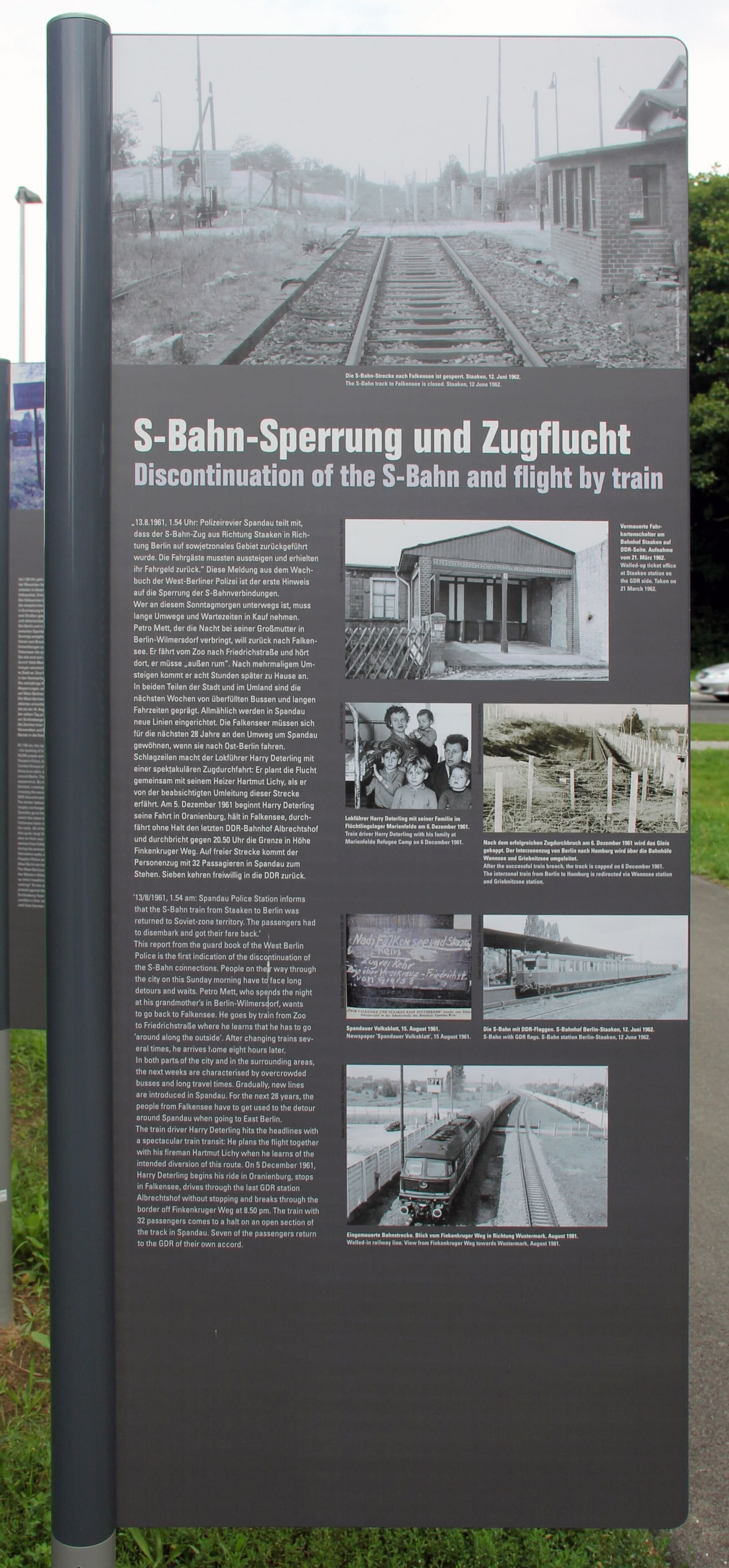 Tall grey outdoor information panel with German and English text and several black-and-white photos, including a family in a refugee camp and a railway line fenced off with posts.