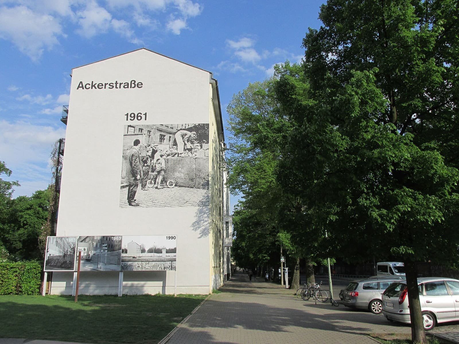 A tall white gable wall reading 'Ackerstraße' and '1961' above a large black-and-white photo of people at a low wall, with a tree-lined street and parked cars to the right.