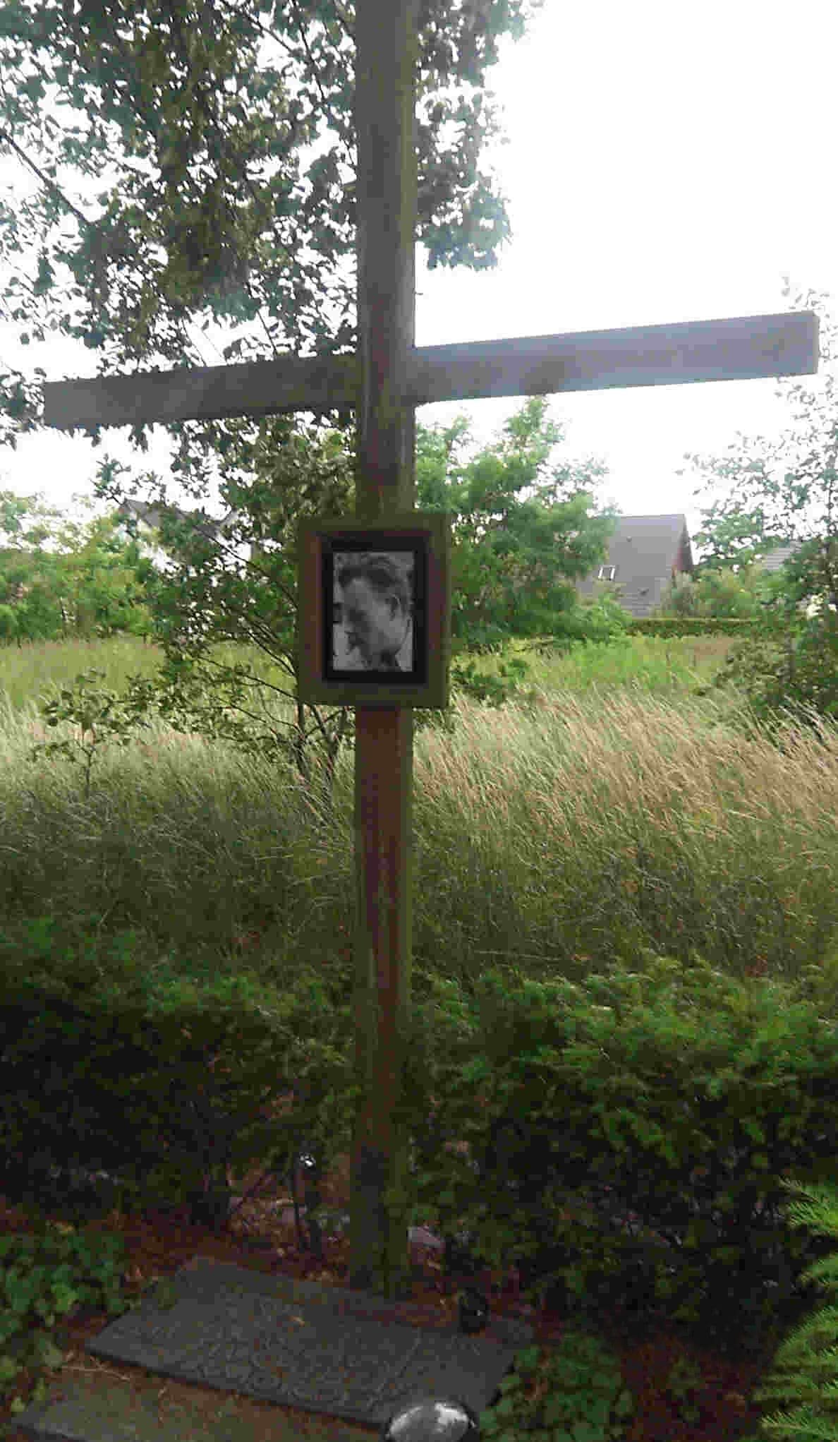 A simple wooden memorial cross with a small plaque, standing on a grassy verge beside a road in a suburban setting