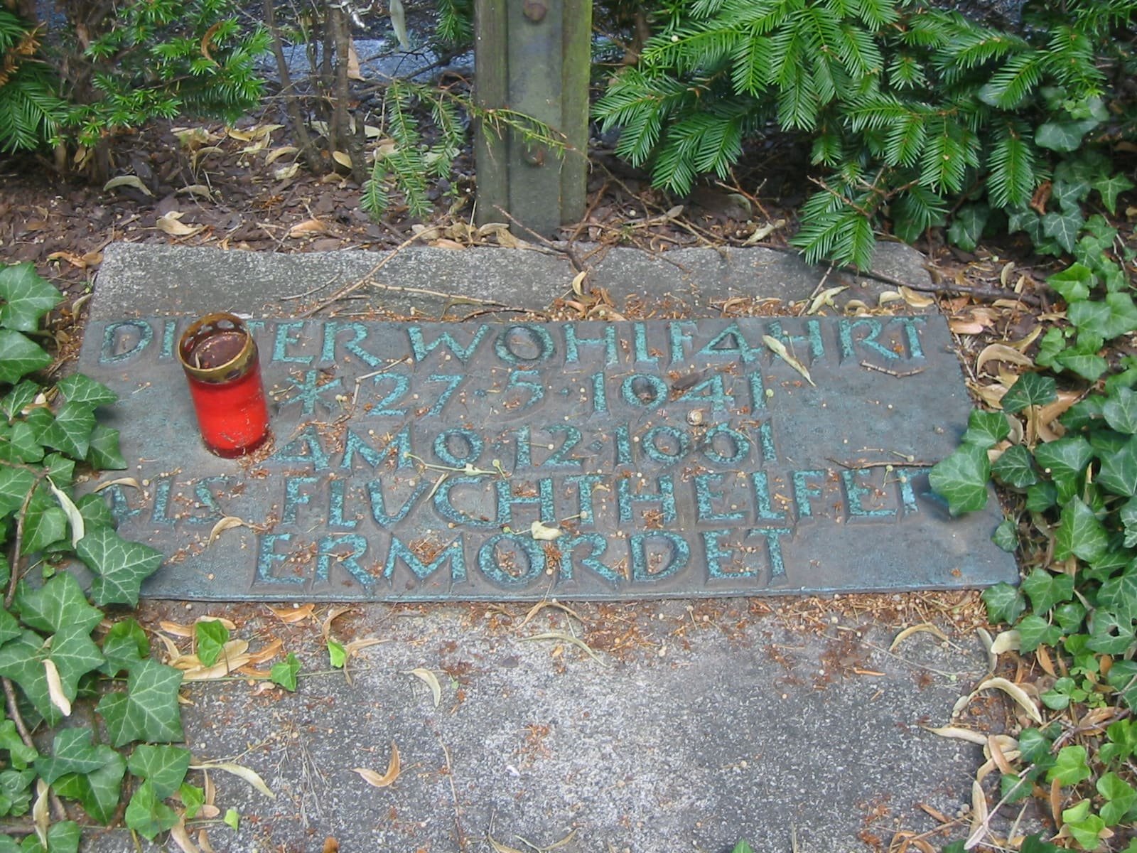 Close-up of a small memorial plaque bearing Dieter Wohlfahrt's name and dates, set at the base of a wooden cross