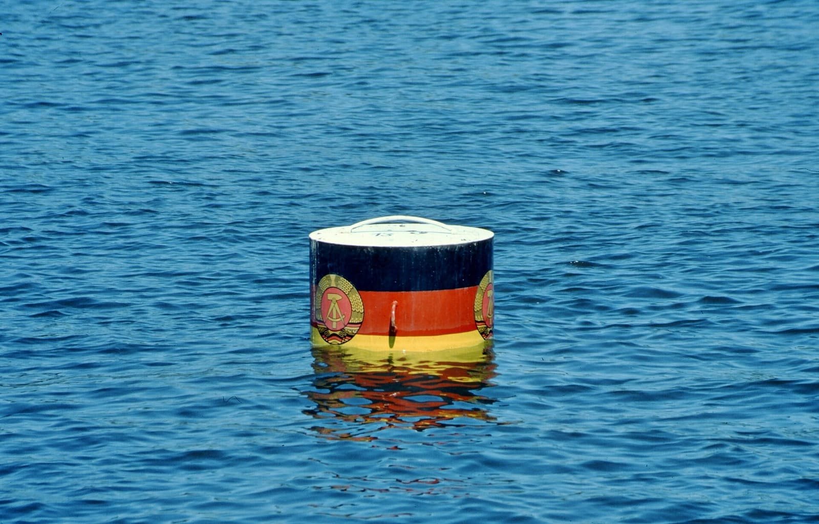 A marker buoy floating on a wide lake, with wooded shoreline in the background.