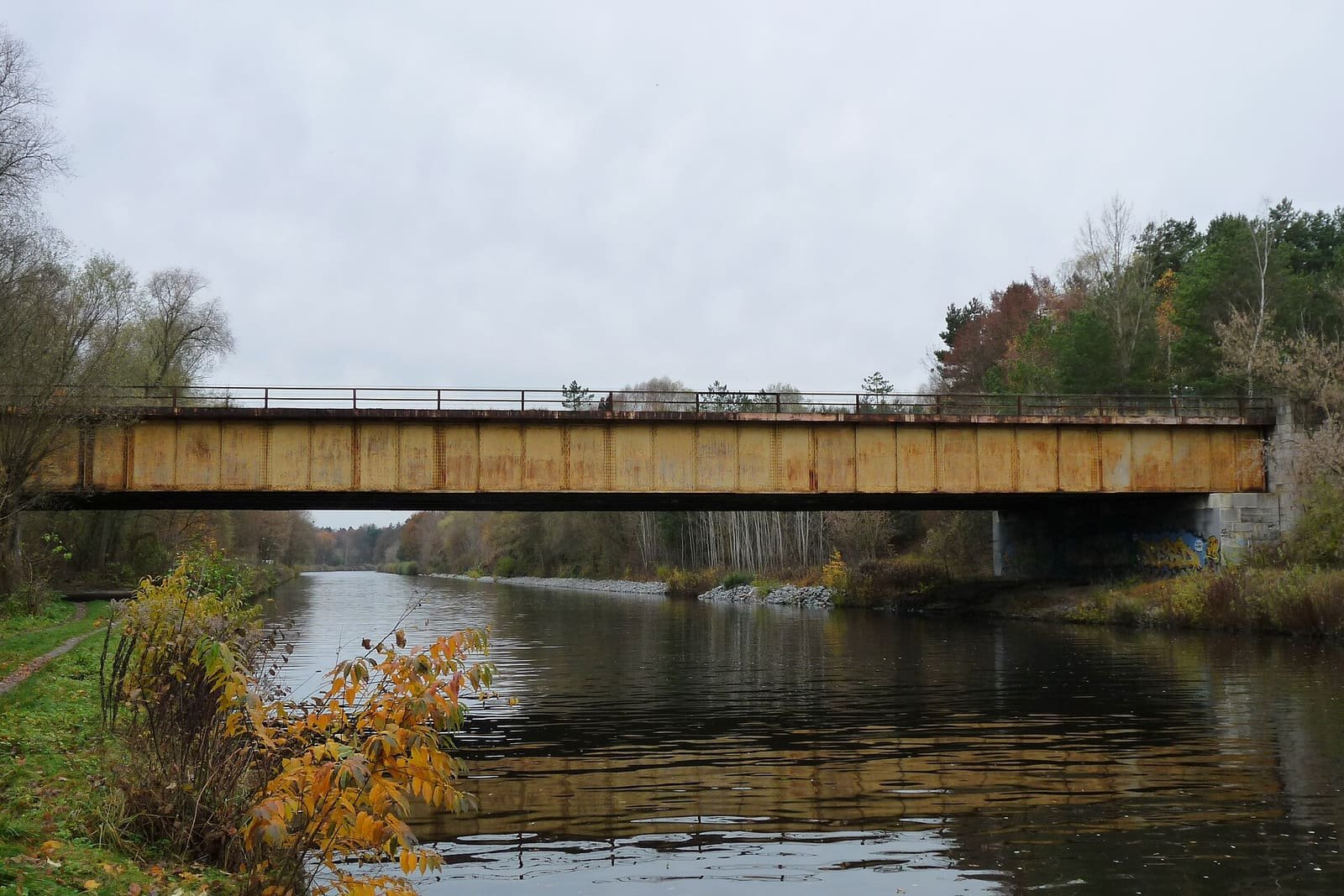 A long rust-coloured girder bridge over a calm canal with autumn trees on both banks.