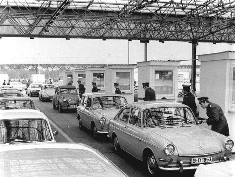 Black-and-white photo: several lanes of 1960s cars, including a Volkswagen estate in front, wait beside small white control booths under a steel-lattice canopy while uniformed officers lean in at the drivers' windows.