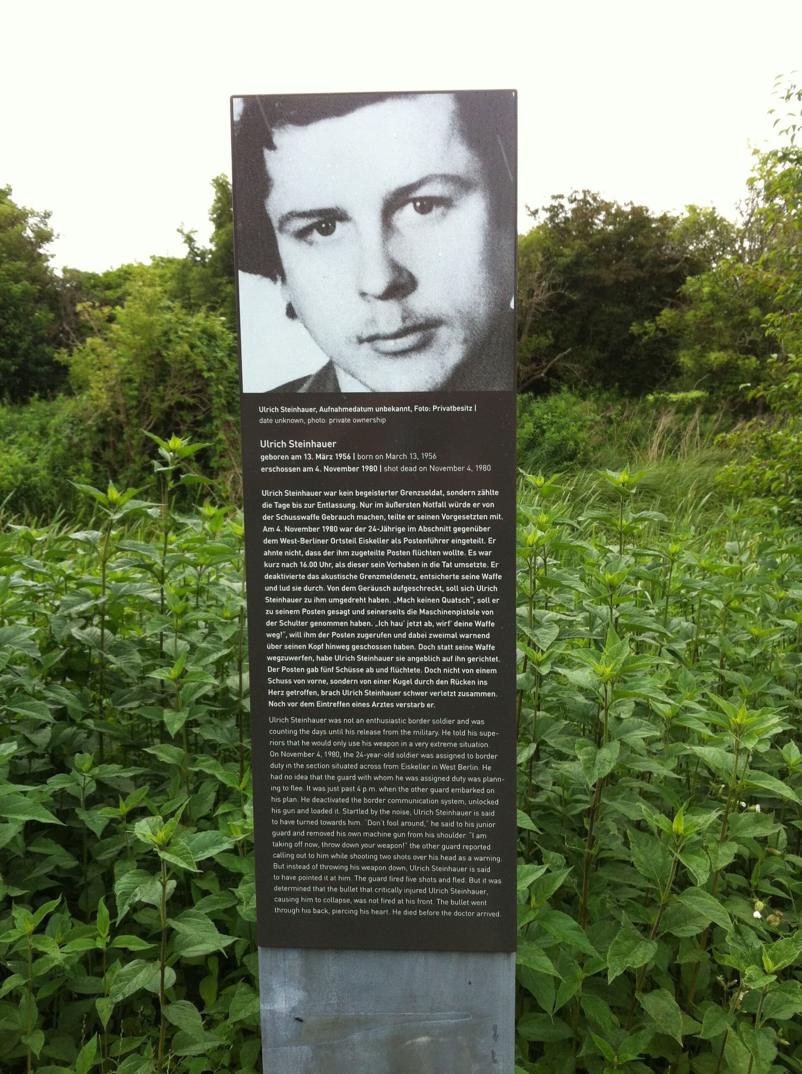 A rust-coloured memorial stele with a portrait and text beside a path at the edge of fields