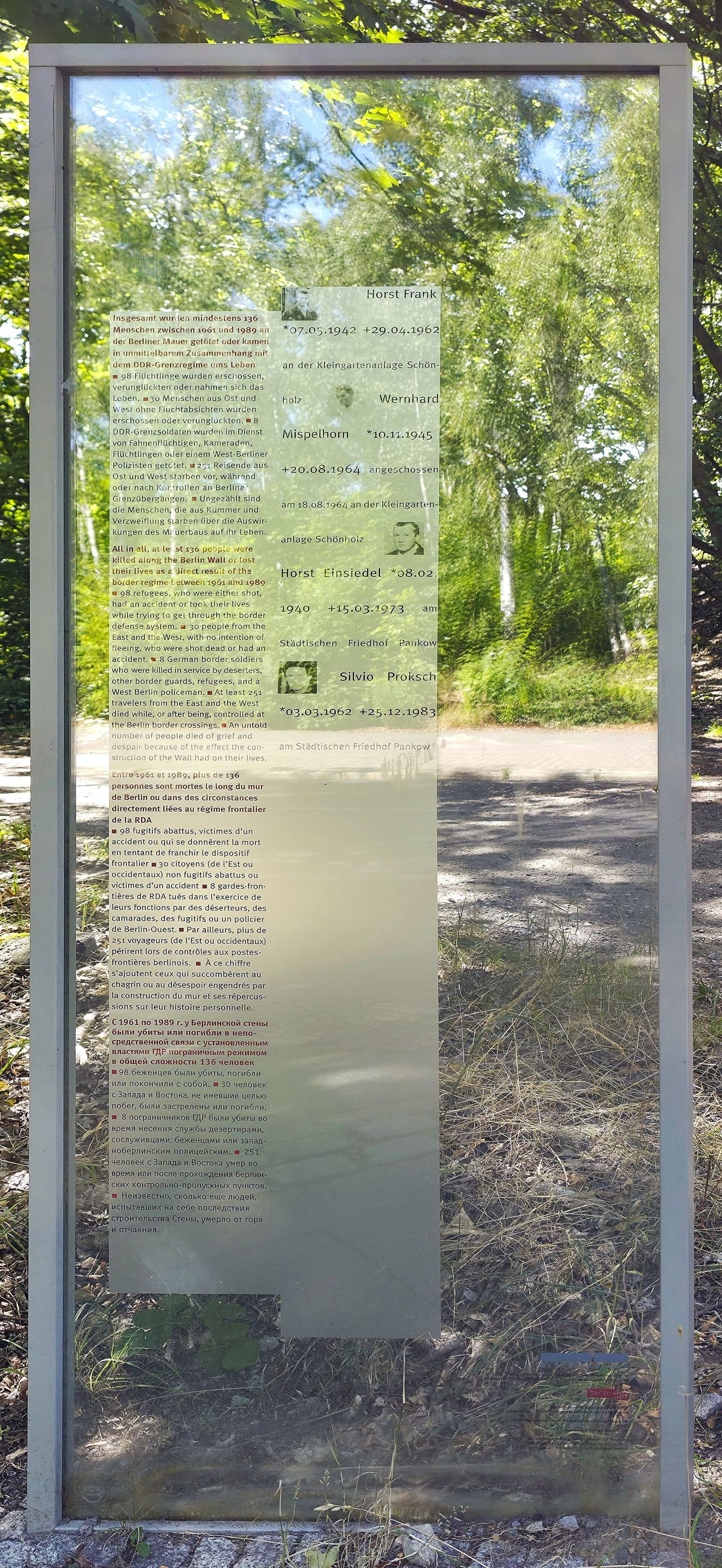 A tall glass panel in a steel frame among trees, printed with text and the names and small portraits of four Wall victims, including Horst Einsiedel.