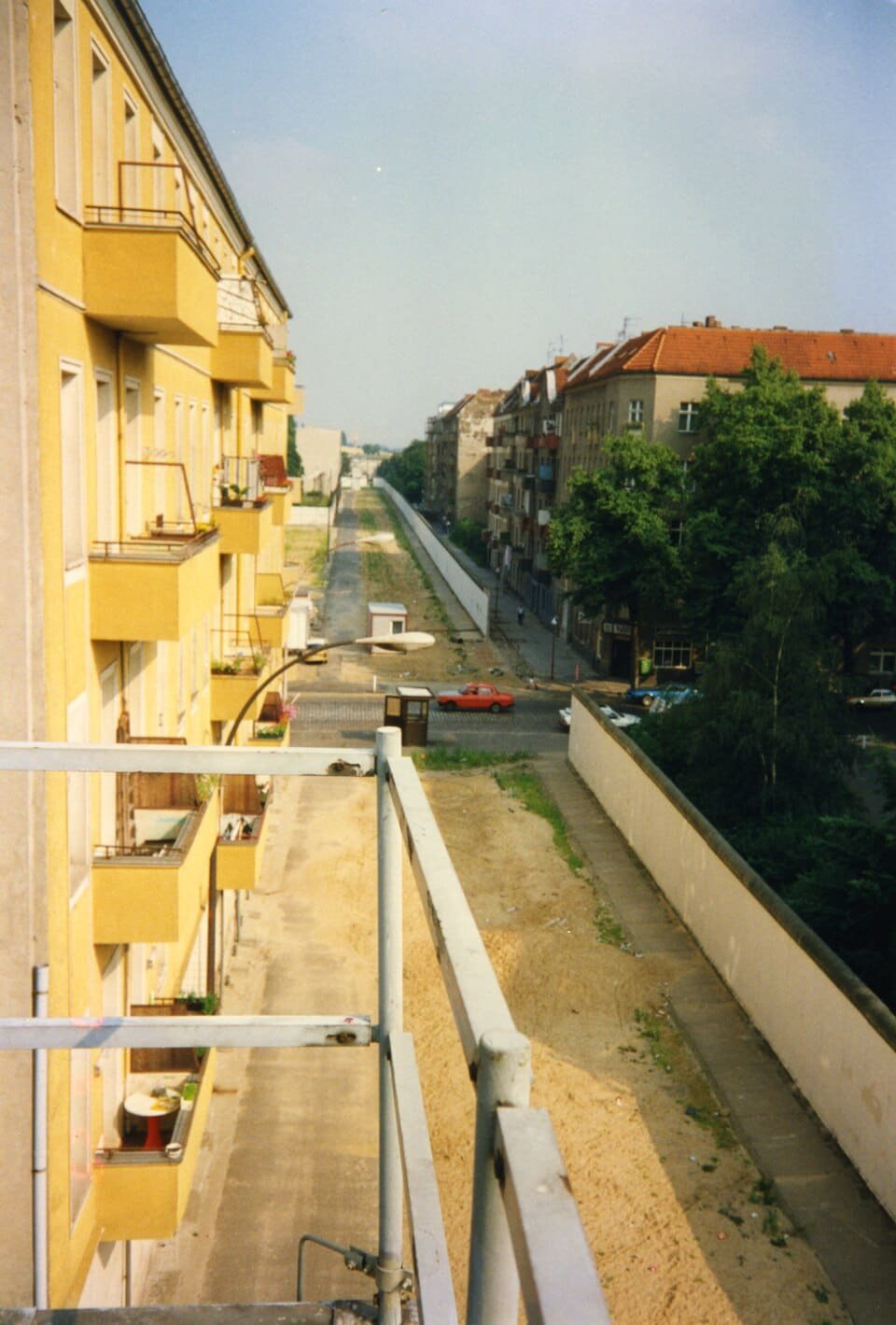 Elevated view along a sandy border strip between a row of yellow apartment balconies and the Wall, with a street and trees beyond.