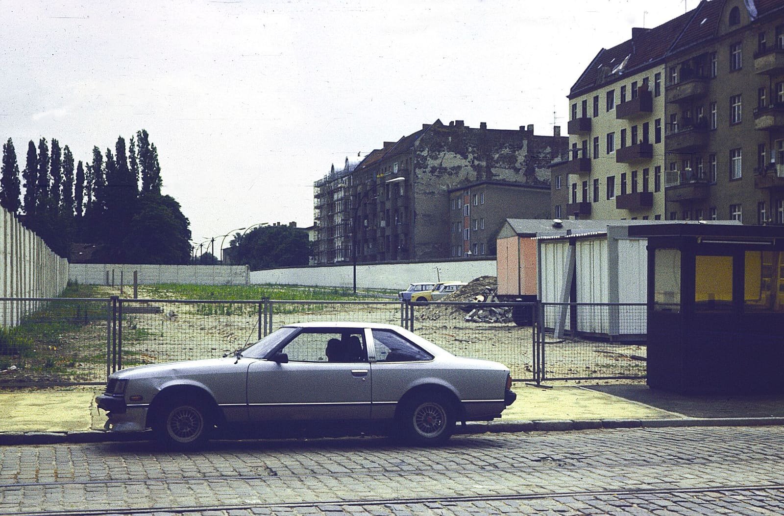 Colour photo of a cobbled street with a parked car, a border booth and a section of the Wall, with old apartment buildings behind.